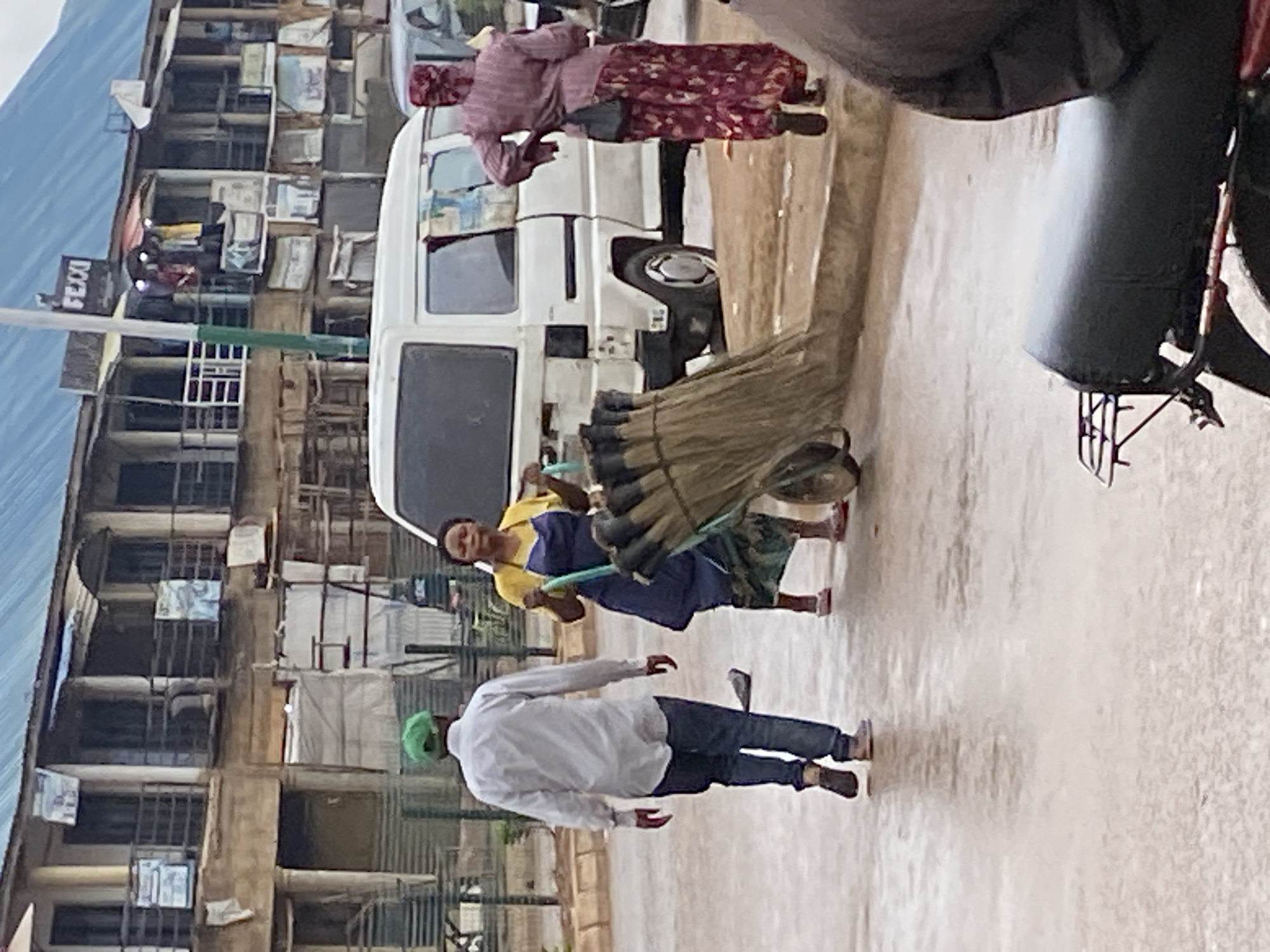 In Bukuru, Nigeria, a street scene unfolds with a white van as a central element. A woman wearing a yellow top and a blue apron pushes a cart laden with brooms to the left of the van. To the left of the van and slightly behind her, a man wearing a white long-sleeved shirt, blue jeans, and a green cap walks away from the viewer. Further to the right, a person in a pink head covering and a patterned pink dress walks away from the viewer, with a dark bag slung over their shoulder. The background features a multi-story building with balconies and various signs. The ground is wet, suggesting recent rain, and the overall lighting indicates an overcast day. In the foreground, the seat of a parked motorcycle is visible, partially obscuring the lower portion of the scene.