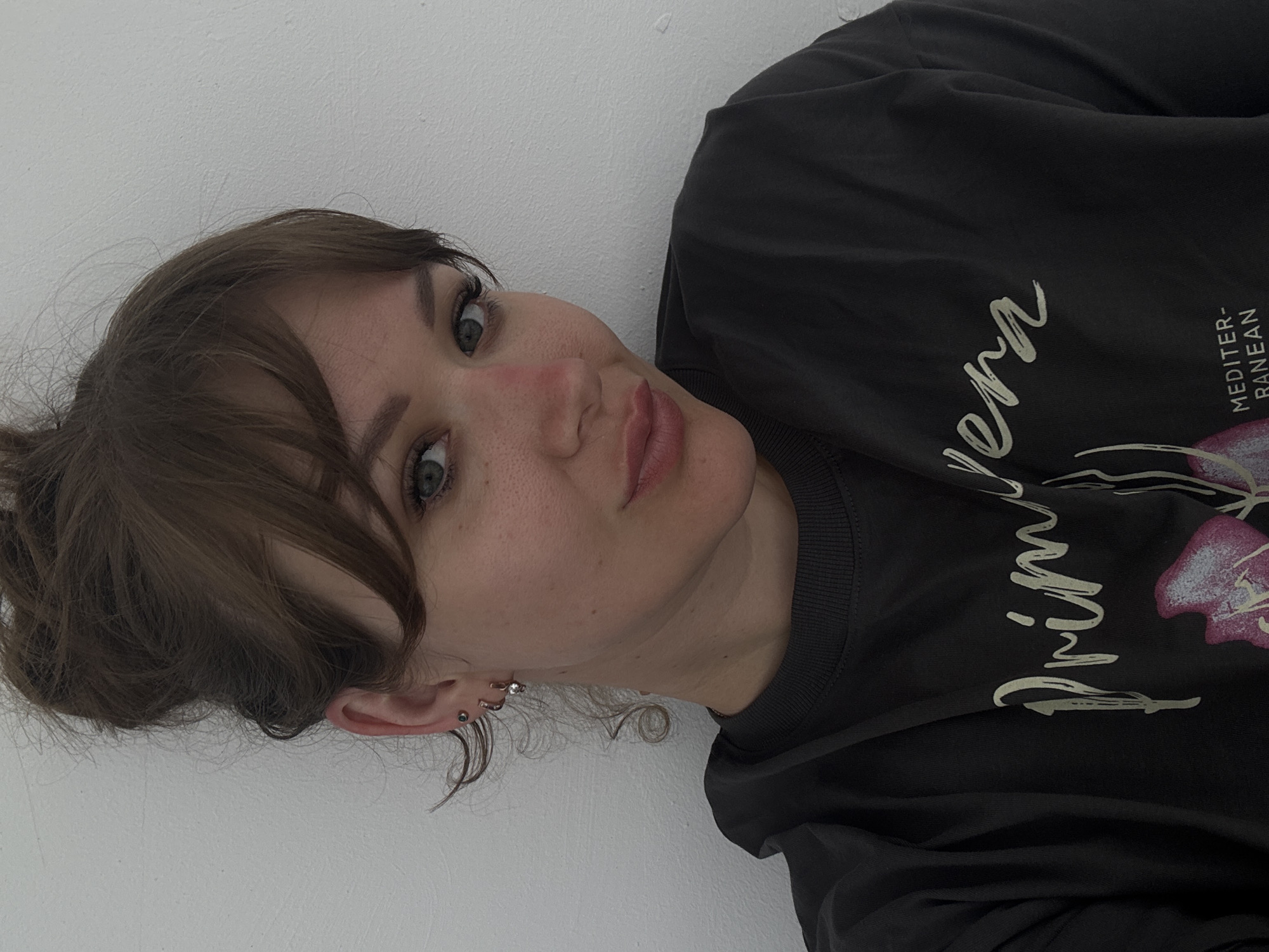 A woman with light skin and brown hair pulled into a bun is positioned against a white wall. She is wearing a gray t-shirt with the word "Primavera" and "MEDITER-RANEAN" printed on it, along with an image of a flower. She has a slight blush on her cheeks and multiple piercings in her ear. The image is taken from a close-up perspective, focusing on her face and upper body. The location is not discernable from the provided image.