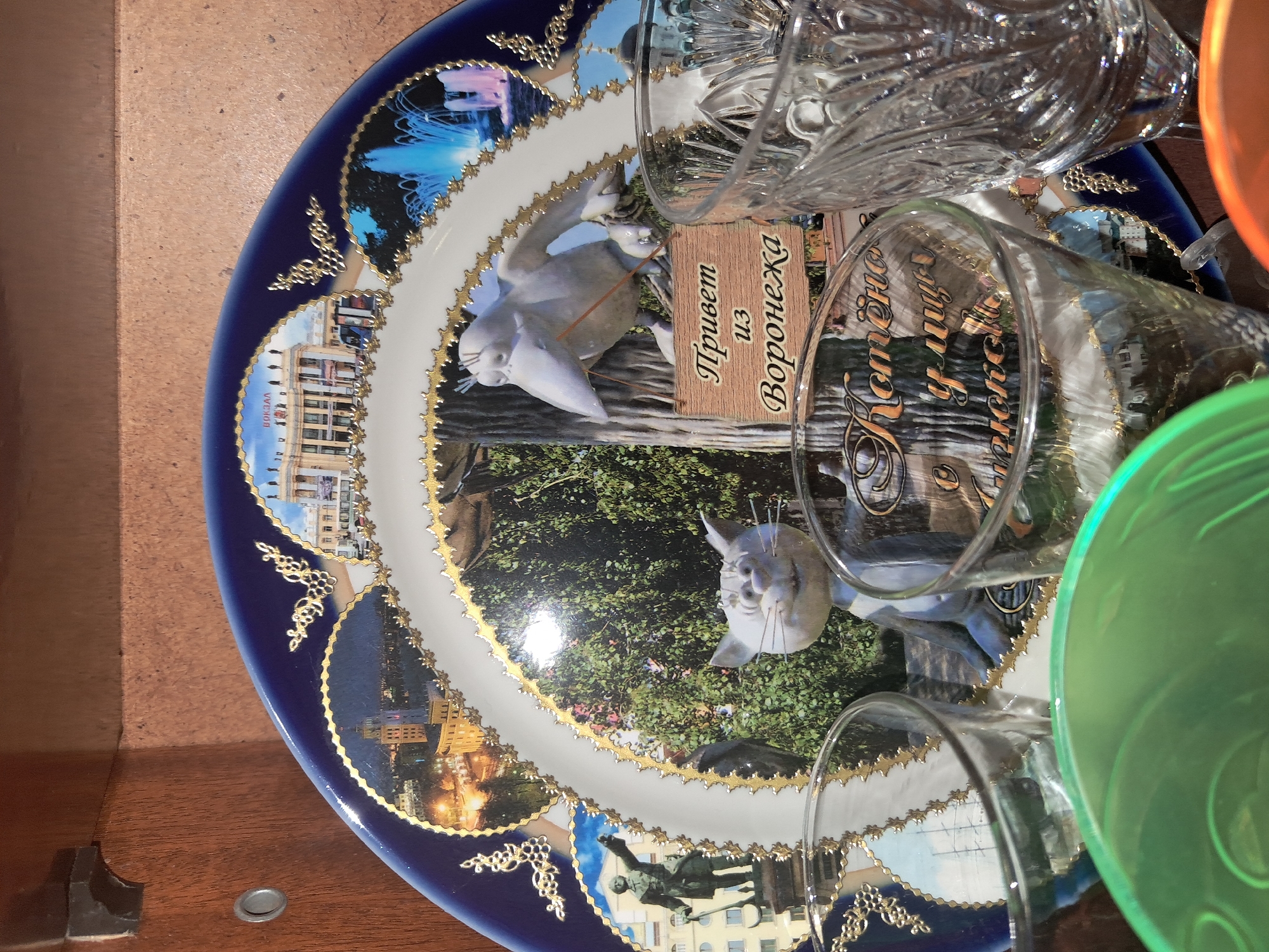 The image shows a decorative plate with various images and text, partially obscured by glassware. The plate has a dark blue border with gold embellishments. The central image features a park-like setting with trees and what appears to be a sculpture of a cat with long whiskers. To the left of the cat sculpture, a bird is depicted with a wooden sign that has Cyrillic text. The text on the sign reads "Привет из Воронежа" (Privet iz Voronezha), which translates to "Greetings from Voronezh." Another section of the plate displays images of buildings, some illuminated as if at night. Another part of the plate shows a statue of a soldier on horseback. The scene is domestic, with glassware placed on top of the plate. No specific activity is taking place, as the objects are stationary. The overall impression is of a souvenir or decorative item from a city in Russia.