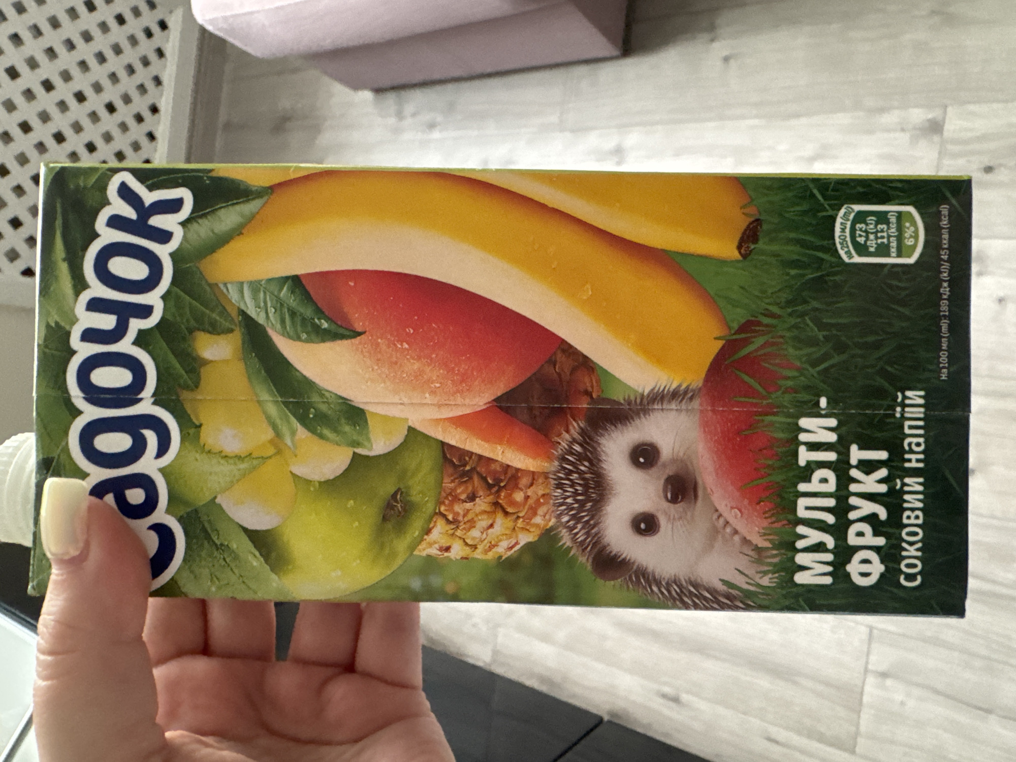 A hand with a light-colored nail is holding a juice box. The juice box has a white cap and features a colorful illustration of various fruits, including apples, peaches, bananas, and a pineapple. There is also a drawing of a hedgehog in the foreground, sitting on green grass. Text on the juice box is in a foreign language, likely Cyrillic script. The background appears to be an indoor setting with a light-colored floor and a blurred pink object on the right. The scene does not provide any information or context to suggest it is located in Tallinn, Estonia.