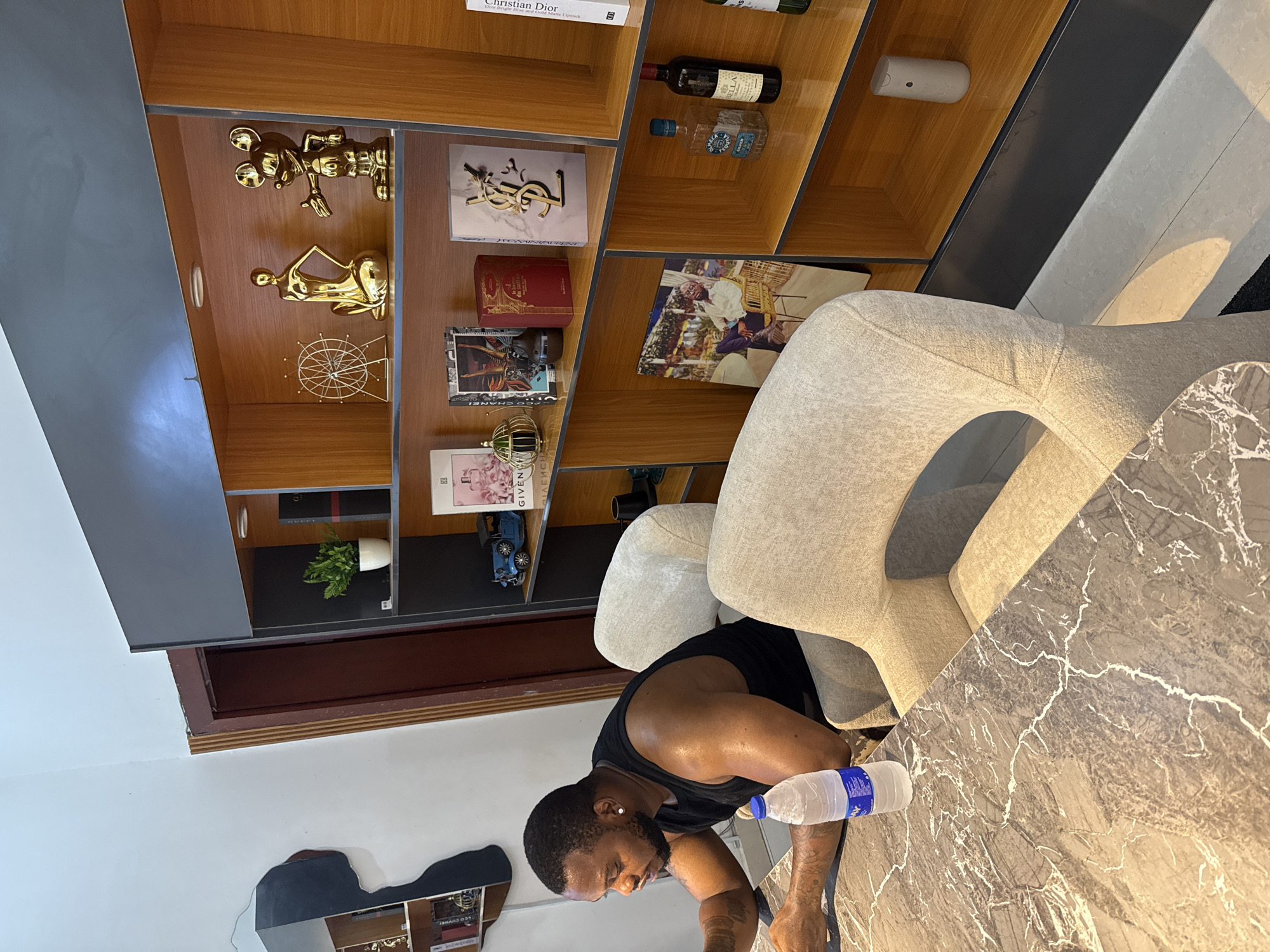 A man sits at a marble table with a water bottle beside him, inside a room featuring a large wooden bookshelf displaying various objects, including figurines, books, and bottles; the room is located in Abuja, Nigeria.