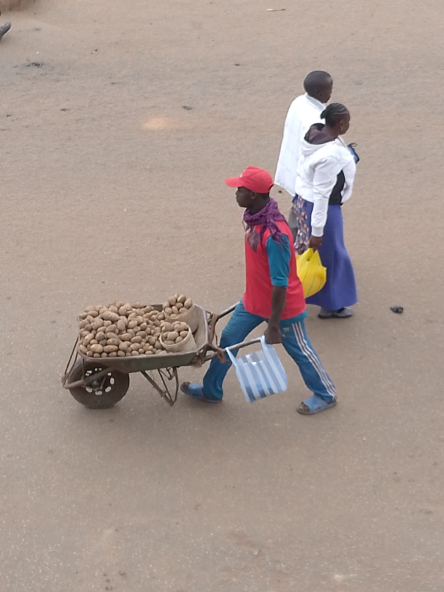 An overhead, full-frame shot shows a sandy, worn street in Bukuru, Nigeria, where three people are visible. In the foreground, a dark-skinned man wearing a red baseball cap, a red t-shirt with teal sleeves, and blue track pants with white stripes, pushes a wheelbarrow filled with potatoes. He wears a purple scarf around his neck and blue sandals. He carries a striped shopping bag in his right hand. Behind him, a woman in a white hooded jacket over a dark garment and a blue skirt walks with a yellow plastic bag. A man in a white robe stands beside her, facing away from the camera. The ground is a muted, dusty brown, with a few scattered dark marks.