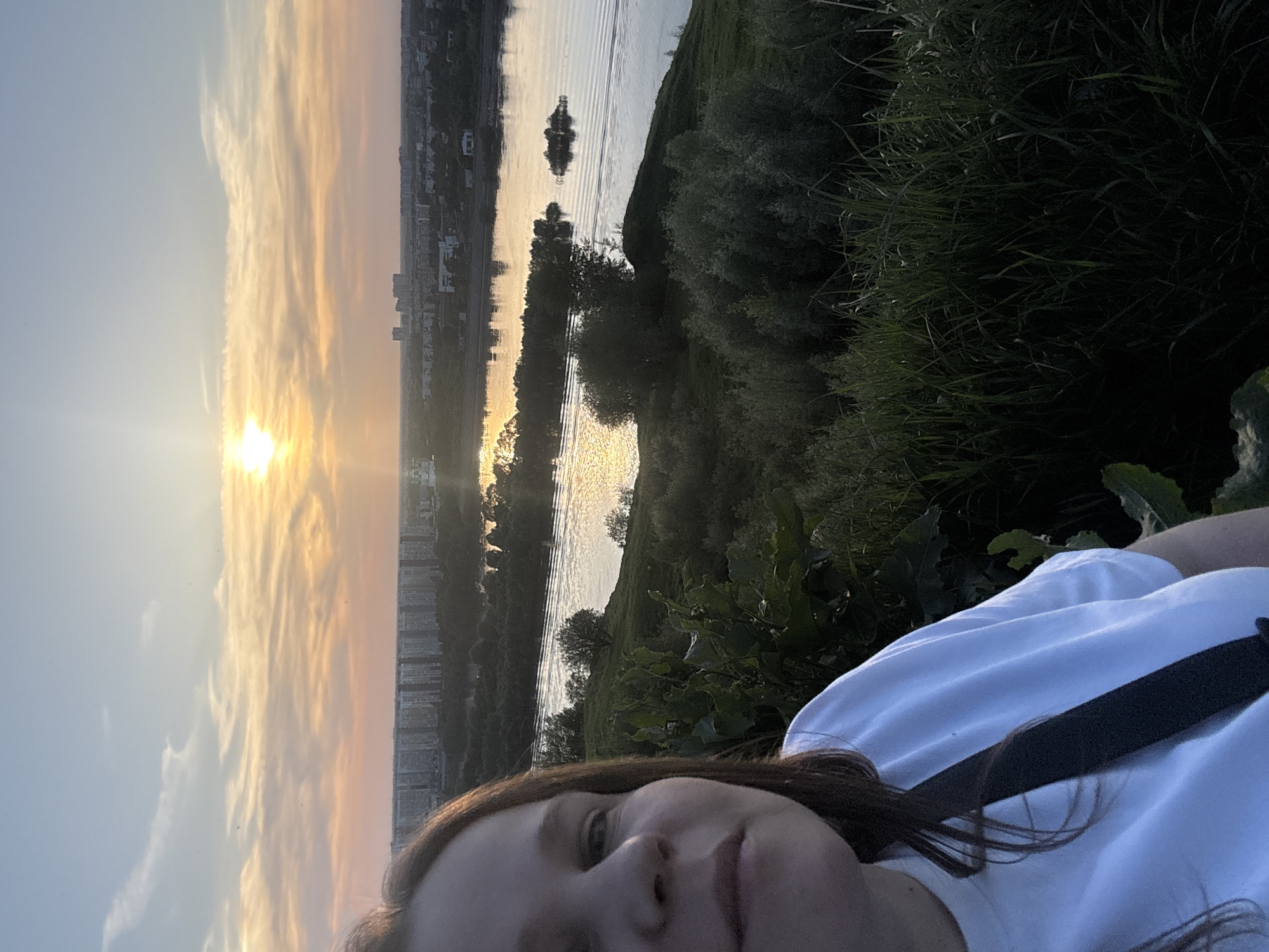A woman is lying on her back in a grassy area overlooking the Oka River in Nizhny Novgorod, Russia. The photo is a selfie, with the woman's face visible in the foreground. She is wearing a white t-shirt and a black strap is across her body. The Oka River is in the midground, with buildings of Nizhny Novgorod visible on the far bank. The setting sun is visible in the sky, casting a warm glow over the scene. The water reflects the light. Greenery, including bushes and tall grass, surround the grassy area where the woman is lying.