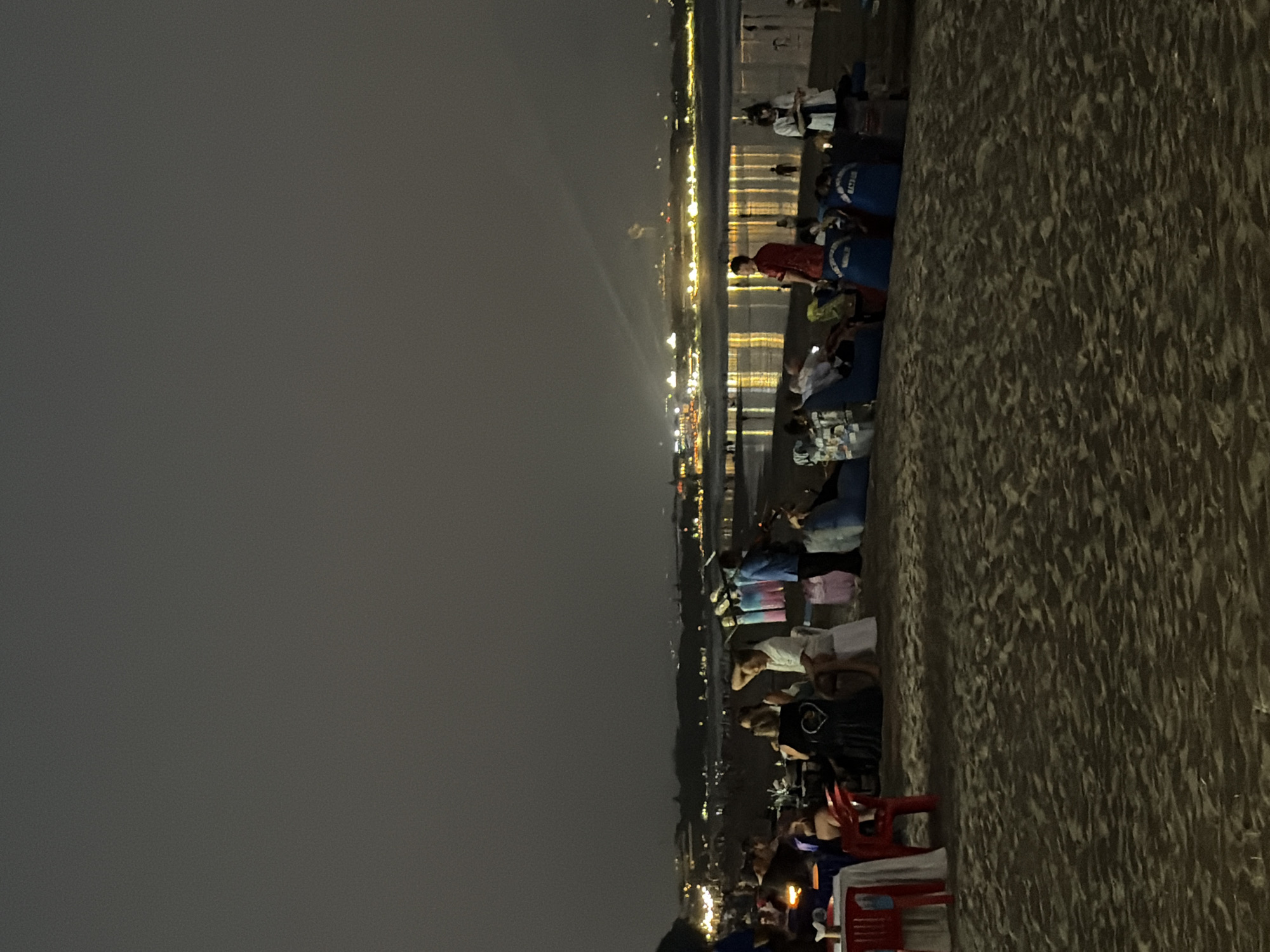 The image depicts a nighttime scene on a beach in Kuta, Indonesia. The sky is dark, suggesting it is evening or night. The beach itself is textured, with sand filling the right side of the frame.
A crowd of people are gathered along the shoreline, facing a body of water that reflects the lights from a distant town or resort area. The lights create a bright, shimmering effect on the water. Many of the people are seated on the sand or on low chairs, indicating a casual gathering or event. Some individuals are standing and observing the scene. The atmosphere appears to be relaxed and communal.
Several objects are visible, including blue bags with white text that reads "BALI WATER SPORTS", suggesting an organized group or activity. There are also red chairs and tables set up, further hinting at a gathering or event. The people are dressed in casual attire.
The overall impression is of a lively beachside gathering at night, possibly a festival, performance, or simply people enjoying the evening by the sea. The bright lights in the distance and the illuminated water create a visually engaging backdrop to the people's activities. There are no clear indicators of specific emotions, but the gathering suggests a sense of togetherness and enjoyment.