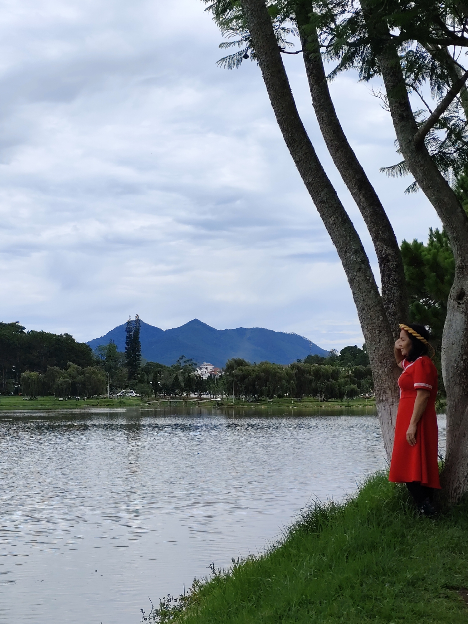 A person stands by trees on the edge of a lake, with mountains in the background. The individual wears a red dress and a straw hat. The location is likely in or near the city of Da Lat, Vietnam. The lake is calm, with reflections visible on its surface. Trees line the distant shore, with the mountains rising beyond. The sky is cloudy.