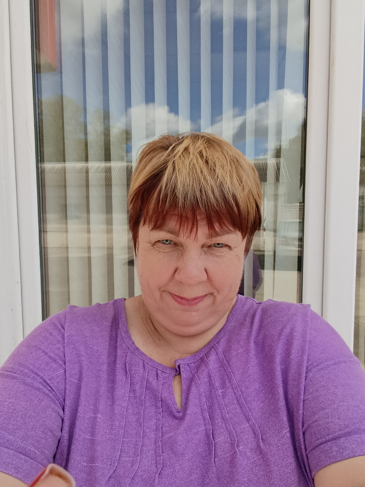 A middle-aged woman with short, layered blonde and auburn hair smiles at the camera in a selfie. She is wearing a purple, short-sleeved shirt with pintuck detailing at the neckline. She is positioned in front of a window with vertical blinds, which reflect the sky with white clouds. The environment appears to be outdoors, possibly on a porch or balcony, given the reflection of trees in the window. The lighting suggests it is daytime. The image was captured in Svietlahorsk, Belarus.