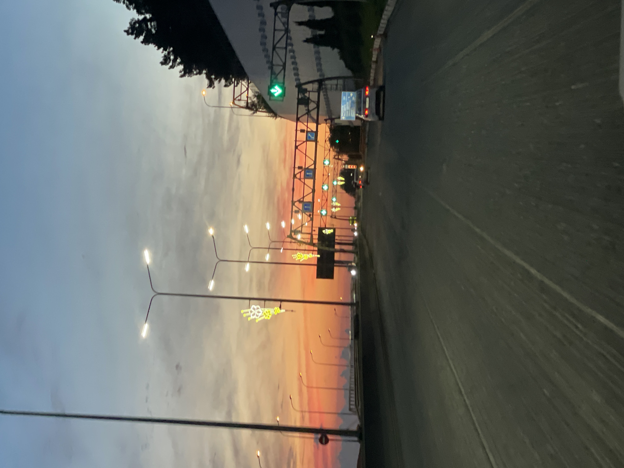 The image depicts a road at sunset in Sochi, Russia, likely near the Adler airport. The sky is a gradient of blue and orange, with scattered clouds. Numerous streetlights and overhead traffic lights illuminate the scene. A white car is visible in the right lane, and other vehicles are further down the road. Trees line the right side of the road, and a white wall with a green directional arrow is prominent. The road appears to be a highway or major thoroughfare, with multiple lanes visible. The overall atmosphere is that of twilight transitioning into night.