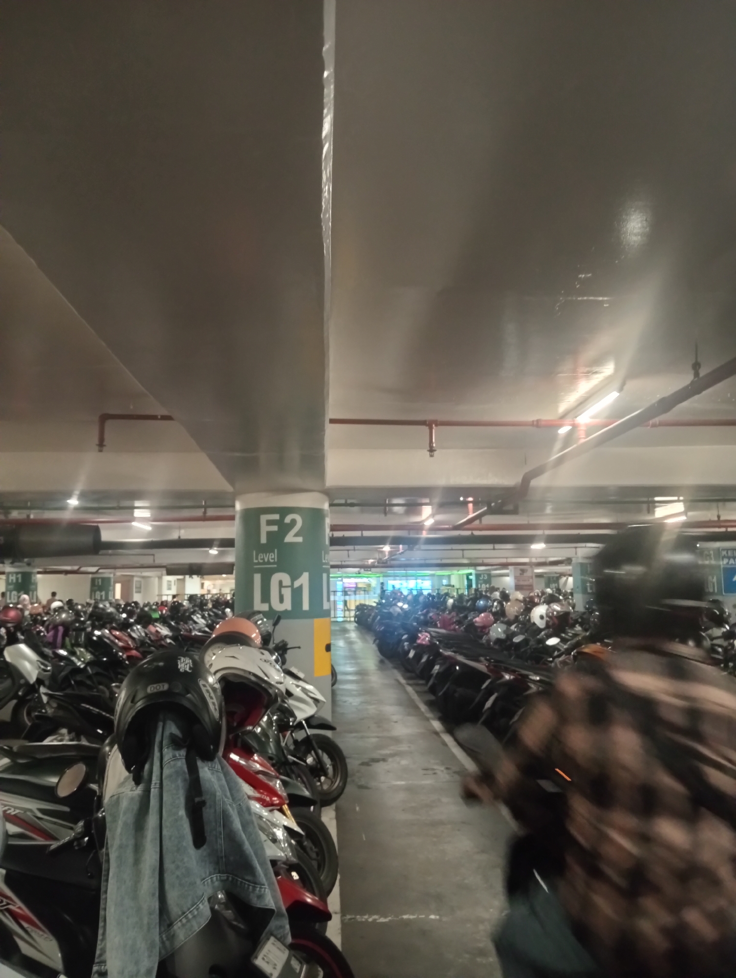 The image depicts a dimly lit indoor parking garage, likely in Surabaya, Indonesia, filled with a dense arrangement of parked motorcycles. A large concrete pillar with signage indicating "F2 Level LG1" is prominently visible on the right side of the frame, marking a section of the parking level. The ceiling of the garage is a dark, concrete structure with exposed pipes and fluorescent lighting fixtures providing illumination.
In the foreground on the left, a motorcycle helmet and a denim jacket are hanging from the handlebars of a parked motorcycle. The jacket is light blue and appears to be a denim jacket. The helmet is black with white lettering. Various other motorcycles are parked in neat rows, stretching deep into the background.
On the right side of the image, a person wearing a patterned shirt and a dark helmet is partially visible, their body blurred by motion. This individual appears to be moving through the parking lot, possibly on a motorcycle that is out of frame or is the same one they are operating. The overall scene suggests a typical day in a busy motorcycle parking facility in a metropolitan area.