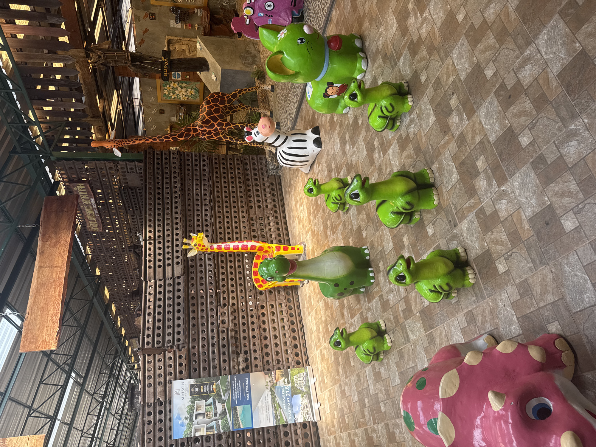 The image shows a collection of colorful animal statues in what appears to be a store or warehouse setting in Ban Khlong Khian, Thailand. The statues include giraffes, zebras, elephants, dinosaurs, and turtle-like creatures. The flooring is tiled with brown and tan square tiles. The statues are arranged on the floor, with the giraffes and a zebra positioned near the middle, along with dinosaurs and turtle-like figures in a line. A pink dinosaur-like statue is at the bottom of the frame. The area above the statues is a metal structure that appears to be the ceiling or roof of a building, with advertisements for "MAISON TROPICAL POOL VILLAS" and "STONE CRISHER".