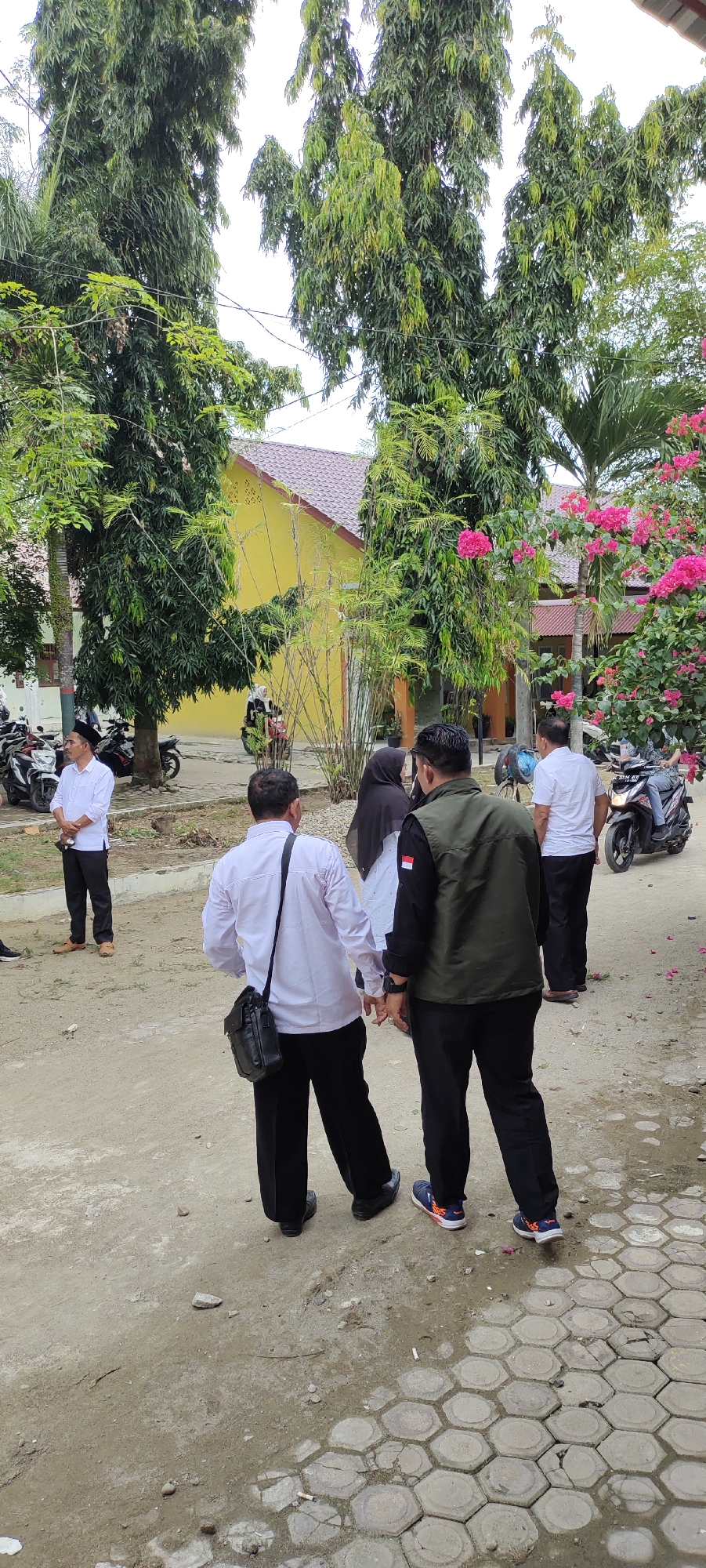 The image depicts an outdoor scene in the city of Meucat, Indonesia, likely within the grounds of a school or administrative building. Several individuals are present, engaged in what appears to be casual conversation or movement. In the foreground, two men are seen from the back. One wears a white long-sleeved shirt and black pants, with a black shoulder bag. The other is dressed in a dark green vest over a black shirt and black pants.
Further into the scene, a man in a white long-sleeved shirt and black pants stands facing left, looking towards a group of parked motorcycles. Behind him, two individuals, one wearing a black hijab, are near a motorcycle. A woman in a black hijab and white outfit is riding a red motorcycle in the middle ground.
The environment features tall, leafy trees and bamboo stalks. A yellow building with a brown tiled roof is visible in the background. The ground is a mix of dirt and paving stones with a hexagonal pattern. Pink flowers are abundant on a bush to the right of the frame. The overall impression is that of a public space, possibly during a daytime event or gathering.