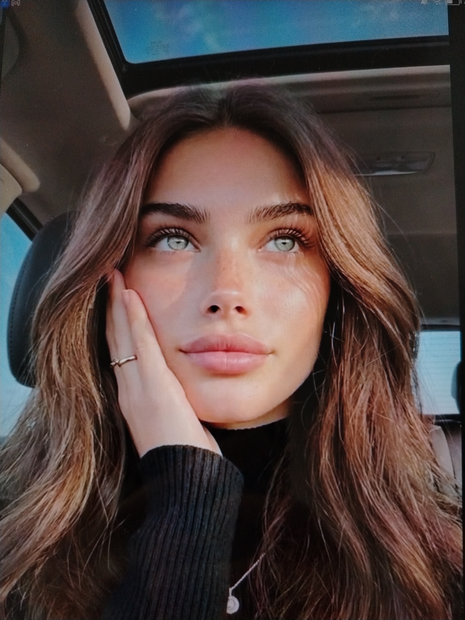 The image is a close-up portrait of a young woman with light skin, long, wavy brown hair, and striking blue eyes. She is looking upwards, with her left hand resting on her cheek. She is wearing a black turtleneck sweater and a thin gold ring on her left ring finger. The background appears to be the interior of a car, with a sunroof visible at the top and a car seat on the left. There is no specific information or visual cues in the image that would indicate the location is Stavropol, Russia. The lighting suggests it might be daytime, possibly with sunlight filtering through the sunroof. The overall impression is one of a casual, candid photograph.
