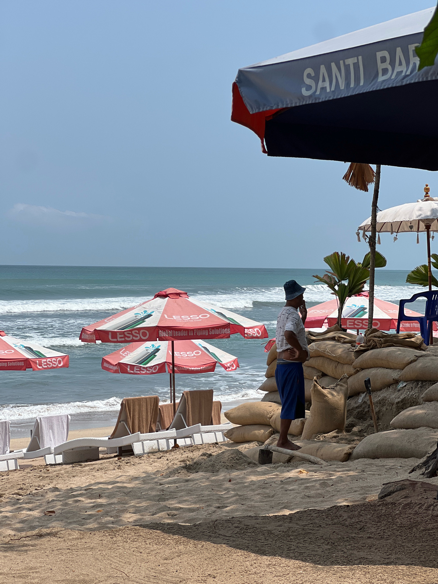The image shows a beach in Kuta, Indonesia. In the foreground, a man wearing a blue hat, white patterned shirt, and blue shorts stands with his hand to his face, appearing to be on his phone. He is positioned behind a barrier made of sandbags. Several red and white umbrellas with "LESSO" branding are visible on the beach, along with white lounge chairs and towels. The ocean with gentle waves is in the background, under a clear blue sky. A large umbrella with "SANTI BAR" written on it is in the upper right portion of the image. The overall scene depicts a beach setting with a barrier under construction or in place.