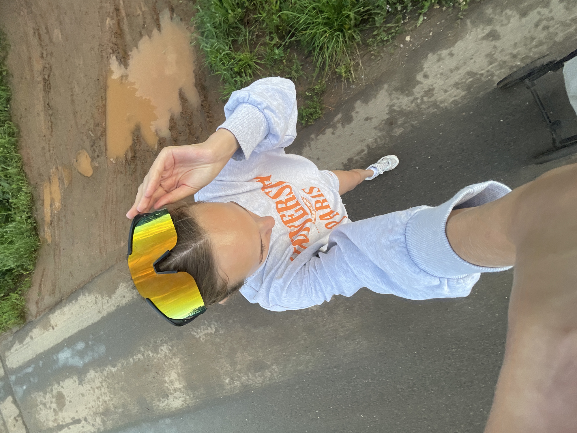 An overhead, wide-angle, first-person perspective image shows a person from the neck down, wearing a light gray long-sleeved sweatshirt with orange text, and white sneakers. The person is wearing large, mirrored wrap-around sunglasses with yellow and green lenses. The person's left arm is extended outwards and upward, holding a phone out of frame to capture the selfie. The person is standing on a wet asphalt surface, with a muddy path and a large puddle to their left. Tall green grass grows along the edge of the muddy path and the asphalt. A dark object, possibly a wheelchair, is visible in the upper right quadrant of the image. The scene suggests an outdoor environment in or near the city of Podshivalovo, Russia.
