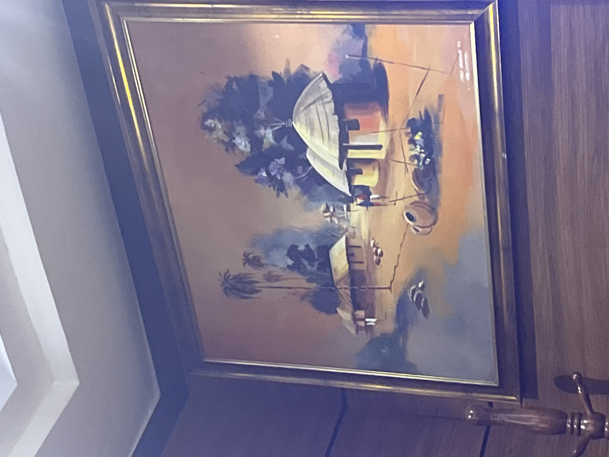 The image displays a framed painting, prominently featuring a vibrant, impressionistic village scene, possibly set in an African landscape. The painting is enclosed within a golden-colored frame and is hung on a textured wooden wall.
Within the painting, the primary subjects are several traditional huts or houses. A large, dome-roofed hut, possibly thatched, stands out in the upper left section, surrounded by dark green and blue foliage, including tall trees. Below it, another, smaller rectangular structure with a similar roof is visible amidst more vegetation and what appears to be palm trees.
The scene depicts daily life in the village. Small, stylized human figures are dotted throughout. One figure is notably positioned near a well-like structure with a pulley system, accompanied by several earthenware pots or vessels, suggesting the activity of fetching water. Other figures are less distinct, seemingly engaged in various everyday tasks. The ground is rendered in earthy tones, transitioning from warmer yellows and browns to cooler blues, implying a distant horizon or body of water. The sky is a blend of warm oranges and blues, indicating a clear daytime setting.
A notable detail is the presence of faint, illegible text or a signature in the upper right corner of the painting. The artistic style uses broad brushstrokes and a rich, warm color palette, conveying a sense of traditional, peaceful village life.
Beyond the painting, a white, angular ceiling or architectural element is visible on the left side of the overall image. In the bottom right corner, a decorative wooden handle with metallic accents can be seen, likely part of a door or furniture item next to the wall where the painting hangs.