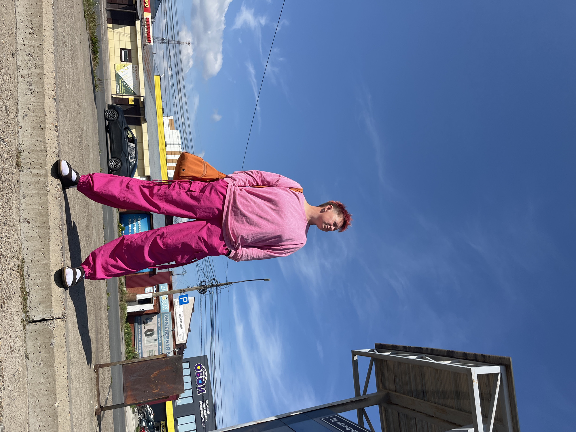 A person with short, pink hair stands on a concrete curb, facing slightly to the right, with their hands in the pockets of bright pink cargo pants. They wear a pink long-sleeved shirt and black and white shoes. An orange sling bag is slung across their body. In the background, a blue sky with wispy clouds stretches above a street scene in Chaikovsky, Russia. Buildings with various signage, including "ОБОИ" (Wallpaper) and "ИНЖЕНЕРНАЯ" (Engineering), line the street. A black car drives on the road, and power lines crisscross the sky. A partially visible bus stop structure stands to the right of the person. The overall scene depicts an urban environment on a sunny day.