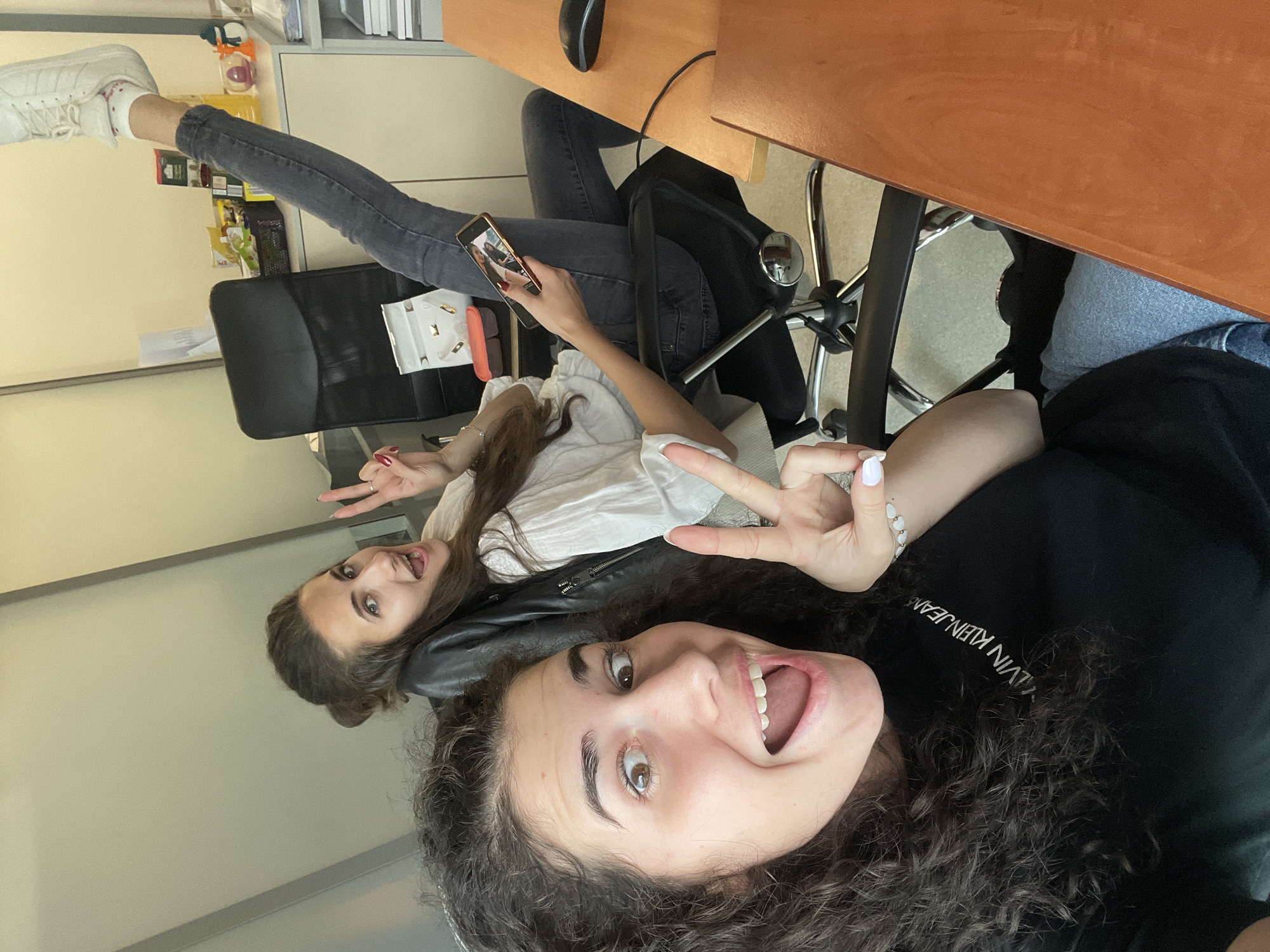 Two young women are posing for a selfie in what appears to be an office setting. The woman in the foreground, wearing a black Calvin Klein jeans shirt, has curly dark hair and is smiling widely with her mouth open, displaying her teeth. She is making a peace sign with her left hand. Behind her, the second woman, wearing a white shirt and a black jacket, is sitting on a chair and also making a peace sign. She is holding a smartphone in her hand, seemingly viewing the image. She is also smiling and has brown hair. One of her legs is raised, with her foot wearing a white sneaker. The office furniture includes a wooden desk with a computer mouse on top and a black office chair. Documents and other office supplies are visible on a shelf in the background. The location is most likely not Sofia, Bulgaria, since there is no information to support this.