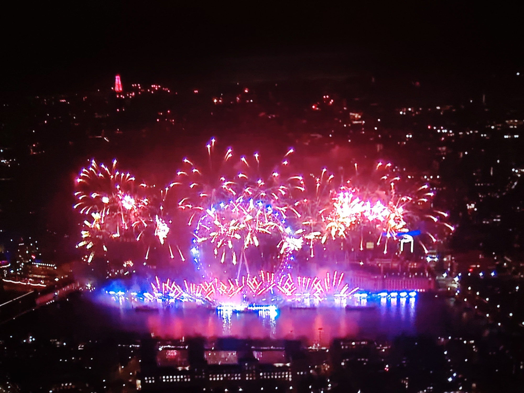 The image depicts a nighttime aerial view of a fireworks display over a body of water. Numerous fireworks are exploding in the sky with bright pink, purple, and white lights, casting reflections on the water below. Along the edge of the water, there are illuminated buildings and structures that appear to be part of a city. In the distance, a tall, illuminated red structure, possibly a monument or tower, is visible against the dark sky. The overall scene suggests a celebration or event taking place in an urban environment at night. The location is identified as Huntingdonshire, United Kingdom.