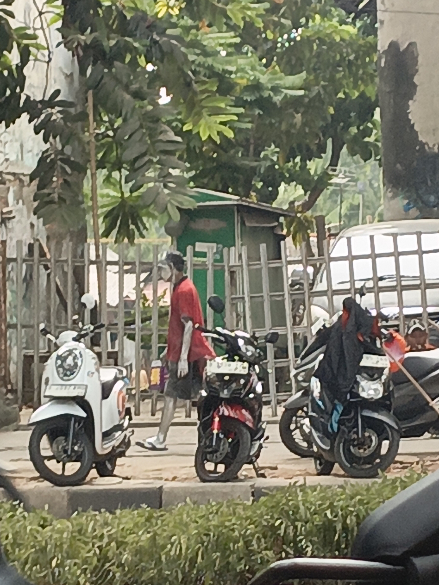 A person wearing a red t-shirt and dark shorts walks past several parked motorcycles. The person is wearing white flip-flops and has a mask pulled down on their face. The scene appears to be on a street in East Jakarta, Indonesia, with a fence, trees, and buildings visible in the background. Two scooters, one white and one red, are parked in the foreground. A dark motorcycle with a black cover is parked to the right of the red scooter. A silver van is partially visible behind the fence.