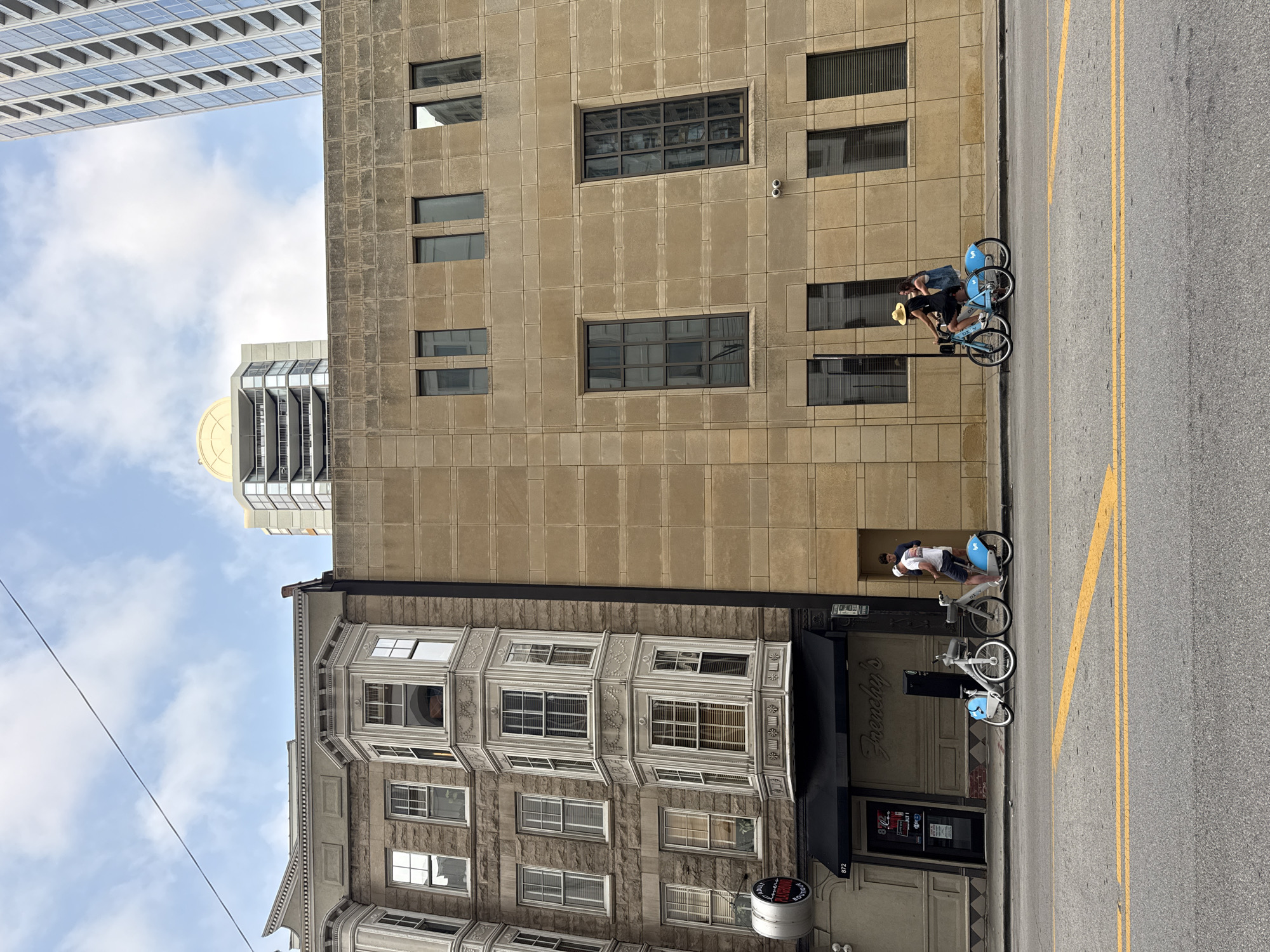 This image depicts a street scene in Chicago, United States of America. On the left is a vintage building with ornate bay windows and stone facade, bearing the signage "Frenchy's". Adjacent to it is a modern building with a tan, tiled facade and rectangular windows. Two people are seen near the vintage building, one standing and the other seated on a blue bicycle. Further to the right, two people are riding a blue bicycle along the sidewalk. The street in the foreground has yellow lane markings. The sky above is partly cloudy.