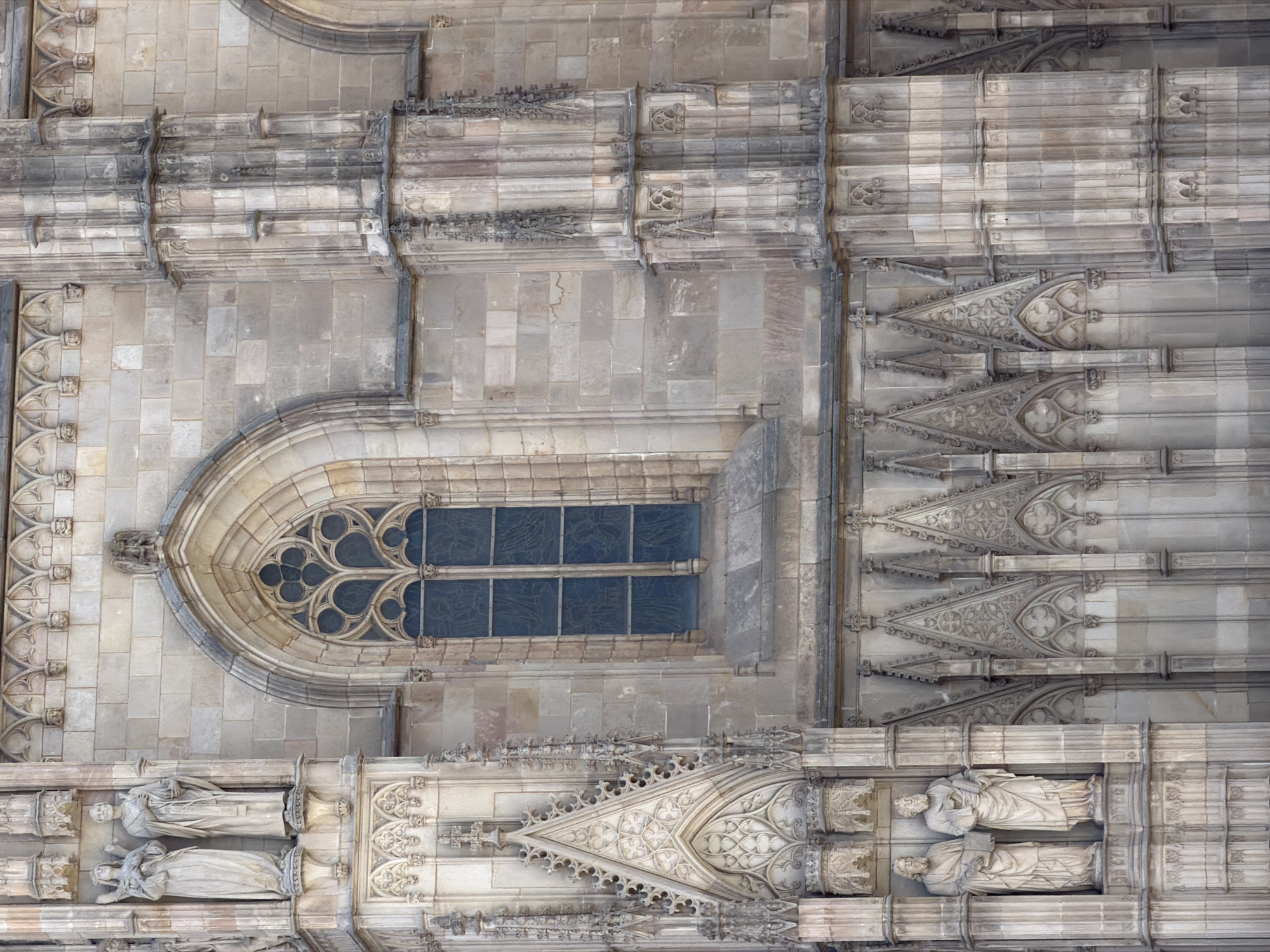 The image shows a close-up, eye-level view of the exterior of the Barcelona Cathedral, located in the Gothic Quarter of Barcelona, Spain. The cathedral's facade is made of light-colored stone blocks and features intricate Gothic architectural details. In the upper left, two full-body stone statues of robed figures stand in niches. To the right of the statues is a large, pointed arch window with ornate tracery, filled with dark blue stained glass. Below this window, the facade is adorned with multiple triangular pediments with elaborate carvings and smaller decorative elements. On the left side of the image, two more full-body stone statues are visible in niches. These figures, also robed, stand on pedestals. The architecture includes vertical columns and detailed decorative friezes running along the building. The overall impression is of a grand, ancient religious structure with a high level of craftsmanship.