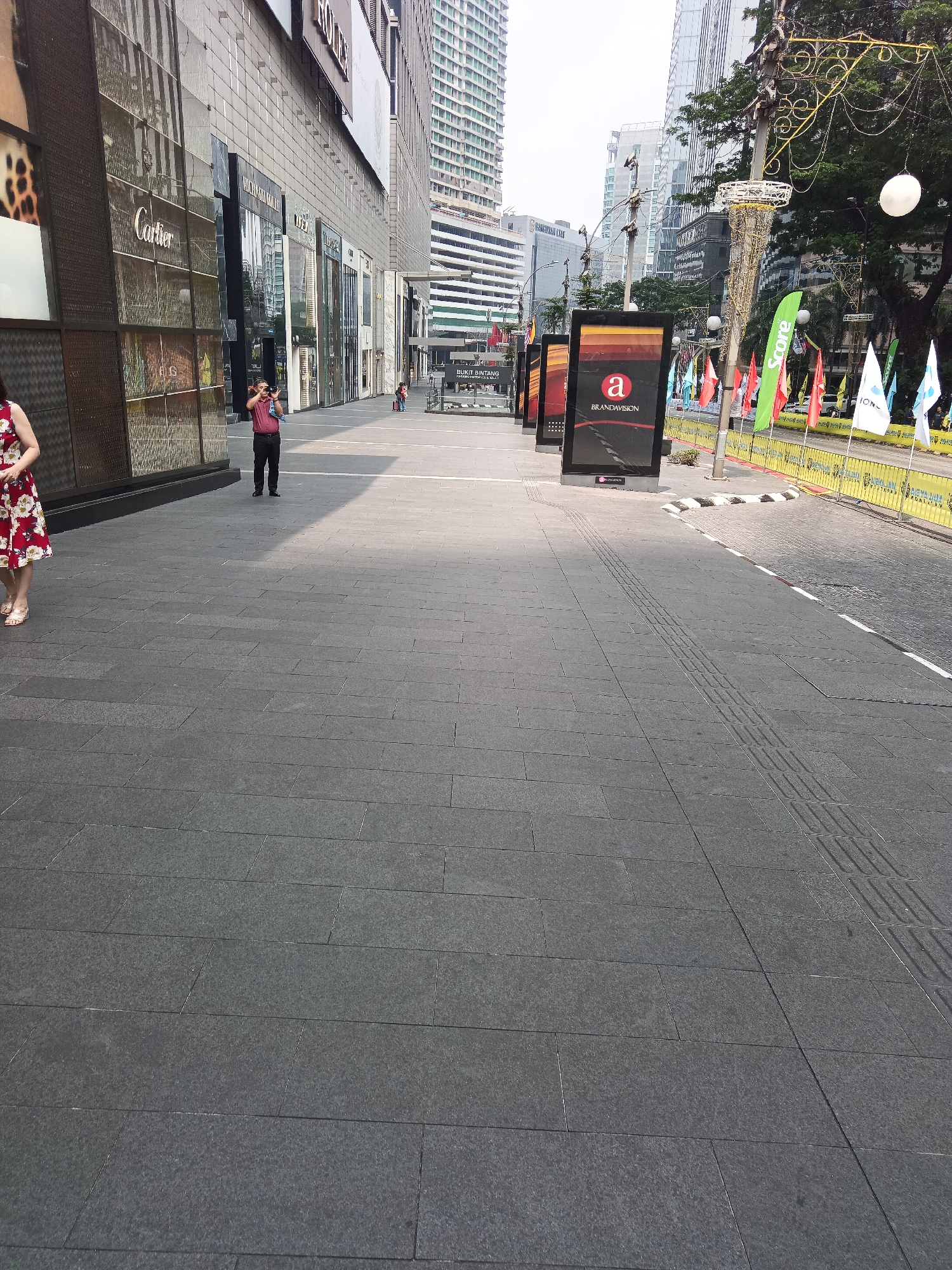 This image depicts a street scene in Kuala Lumpur, Malaysia, likely in the Bukit Bintang area, known for its upscale shopping and entertainment. The foreground is dominated by a wide, gray tiled pedestrian walkway. To the left, the facade of a modern building with large glass windows, displaying store names such as Cartier and Rolex, forms the backdrop. A woman in a red floral dress is partially visible on the far left, walking away from the camera. In the middle ground, a man in a red shirt and black pants is standing, holding a camera and appearing to take a photo. A row of digital advertising screens stands along the right side of the walkway. Beyond the screens, a street is visible with yellow barriers, flags, and trees. The background features tall, modern skyscrapers, characteristic of Kuala Lumpur's urban landscape. The lighting suggests it is daytime, with sunlight casting shadows on the walkway.