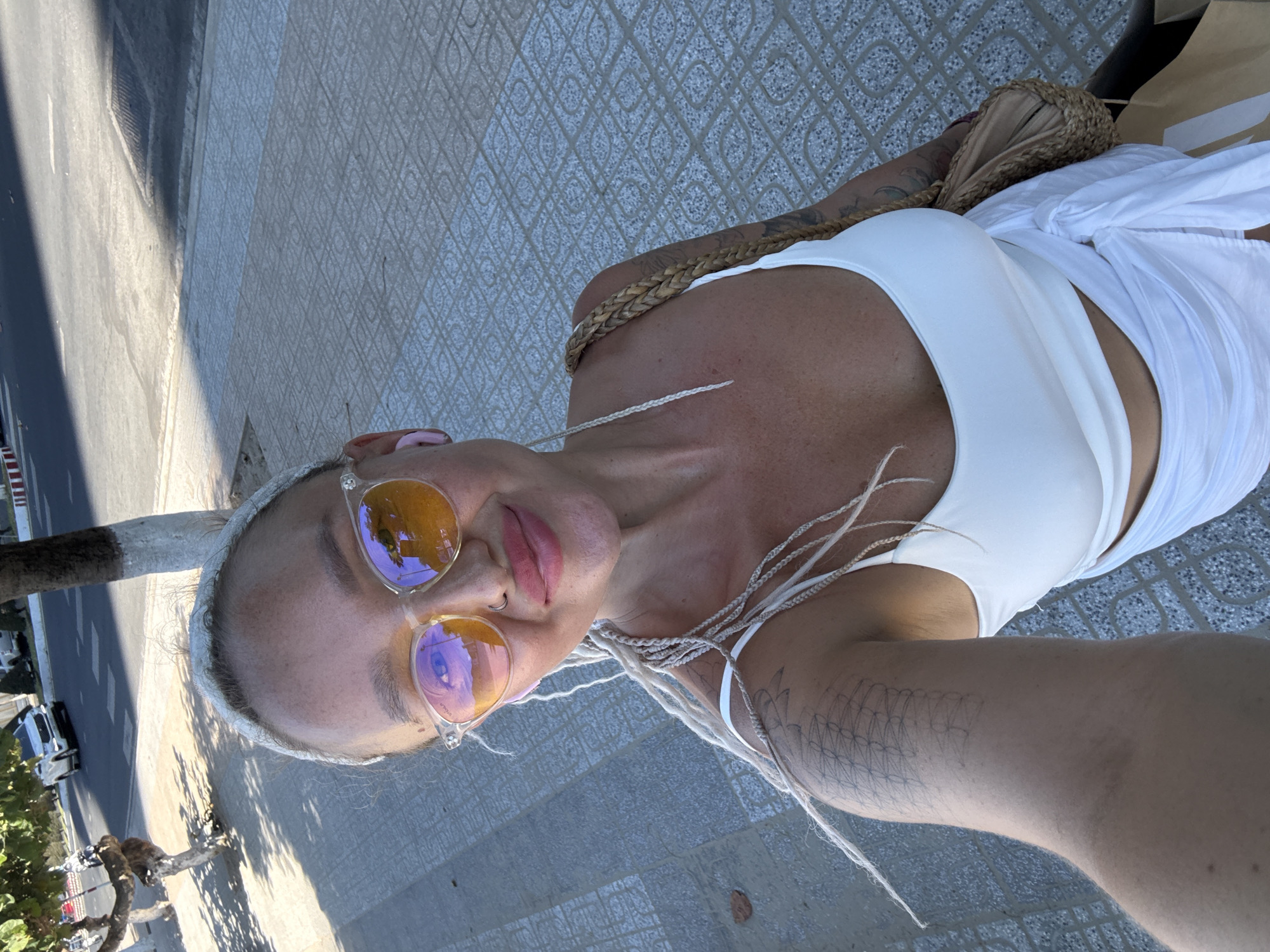 A woman with blonde braided hair and orange-tinted sunglasses is taking a selfie outdoors. She is wearing a white tank top and a white sarong. Her left arm, visible in the foreground, has a tattoo. She is standing on a sidewalk made of patterned tiles. In the background, a road with traffic and trees can be seen. The scene appears to be in a city, possibly near a beach given the light clothing and sunny weather. The specific location is indicated as Bac Nha Trang Ward, Vietnam.