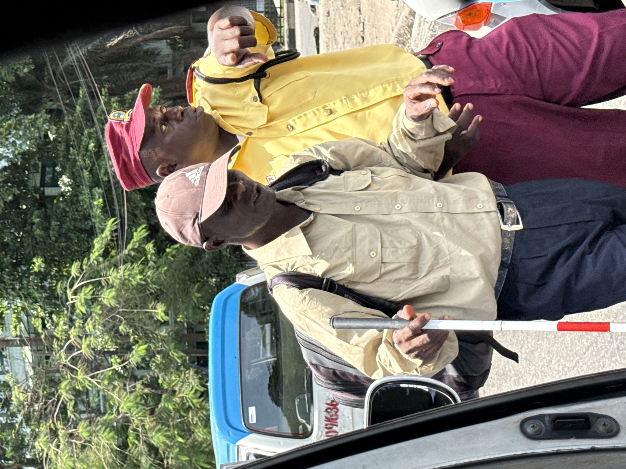Two men stand near a vehicle. The man closest to the camera wears a light brown baseball cap, a light brown collared shirt, and dark blue pants. He holds a white cane with a red stripe. The other man wears a red baseball cap, a yellow collared shirt, and maroon pants. The scene is in Lagos, Nigeria, and the vehicle number is 091636. Trees are visible in the background.