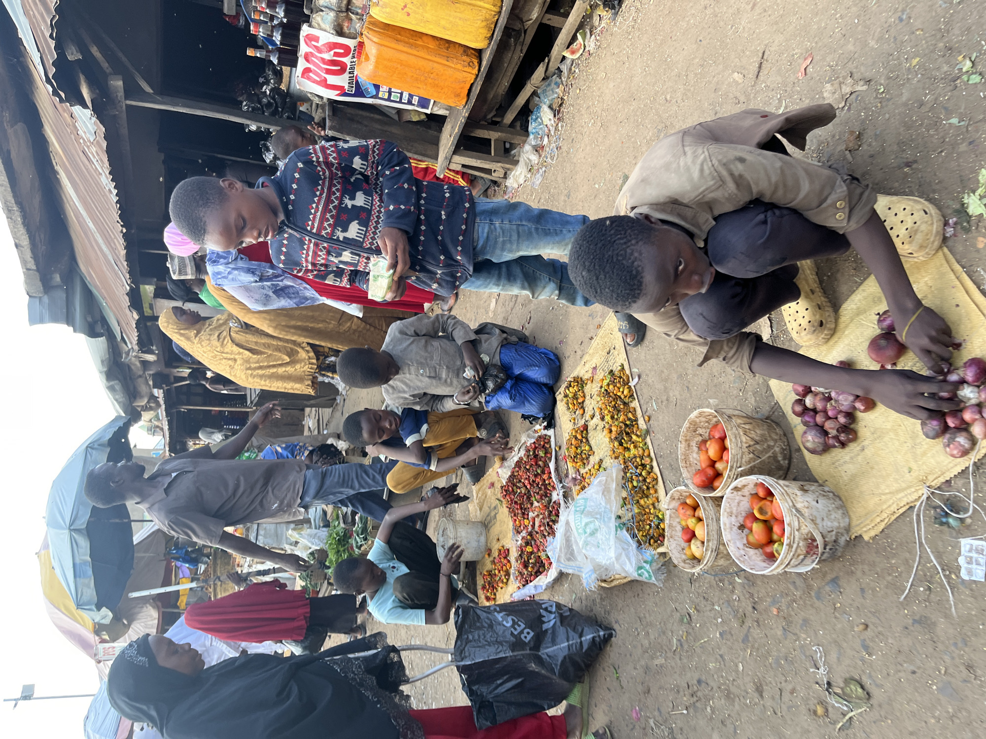 This is a street market scene in Rafin Par, Nigeria. Several individuals, primarily children, are engaged in the sale of produce. In the foreground, a young boy in a light brown jacket and dark pants is crouched on a yellow mat, sorting through a pile of red onions. He is wearing large yellow clogs. To his right, another boy in a blue and white striped shirt and yellow pants is reaching towards a pile of red and yellow peppers. Behind him, a boy in a grey shirt and blue pants is sitting and looking at his hands. In the middle ground, a group of people are gathered around mats spread with various types of peppers. One boy in a blue and red patterned jacket is holding some papers, possibly money, and looking down. A woman in a black hijab and a woman in a red veil are visible on the left side of the image. A man in a brown shirt and blue jeans is walking past. The market stalls have corrugated metal roofs, and various items like bottles of liquid and large yellow containers are displayed. The ground is dusty and appears to be unpaved. Buckets filled with tomatoes are also visible.