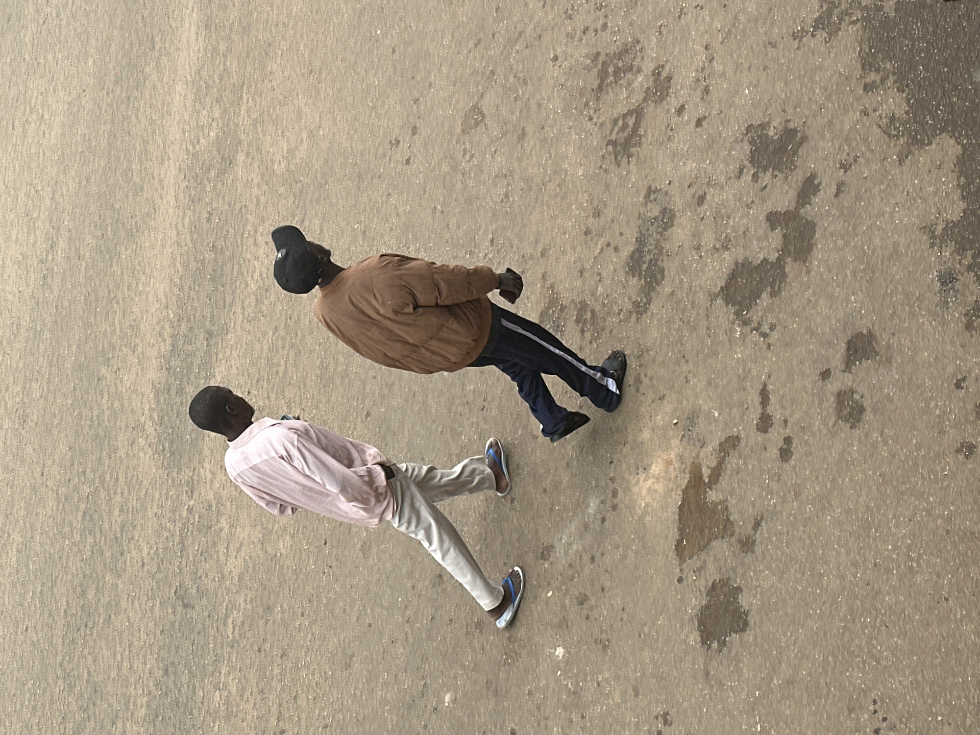 In Bukuru, Nigeria, two individuals are captured in a high-angle, full-body shot walking on a textured, asphalt-like surface. The ground is marked with irregular dark patches, possibly from oil or water stains, scattered across the predominantly light brown terrain.
The person on the left, an adult male, is wearing a pale pink long-sleeved shirt and light-colored trousers. He has his hands in his pockets and is wearing blue and white flip-flops. His body is positioned with his left leg forward, indicating forward motion.
To the right, another adult male walks with his body angled slightly away from the camera. He is wearing a brown puffer jacket over a dark long-sleeved shirt and dark pants with a white stripe down the side. He has a black baseball cap on and is wearing dark slip-on shoes. His right hand is visible, holding a dark rectangular object, possibly a phone. His left leg is forward, also suggesting movement. The two individuals appear to be walking in the same direction, with the one on the right slightly ahead and to the right of the one on the left.