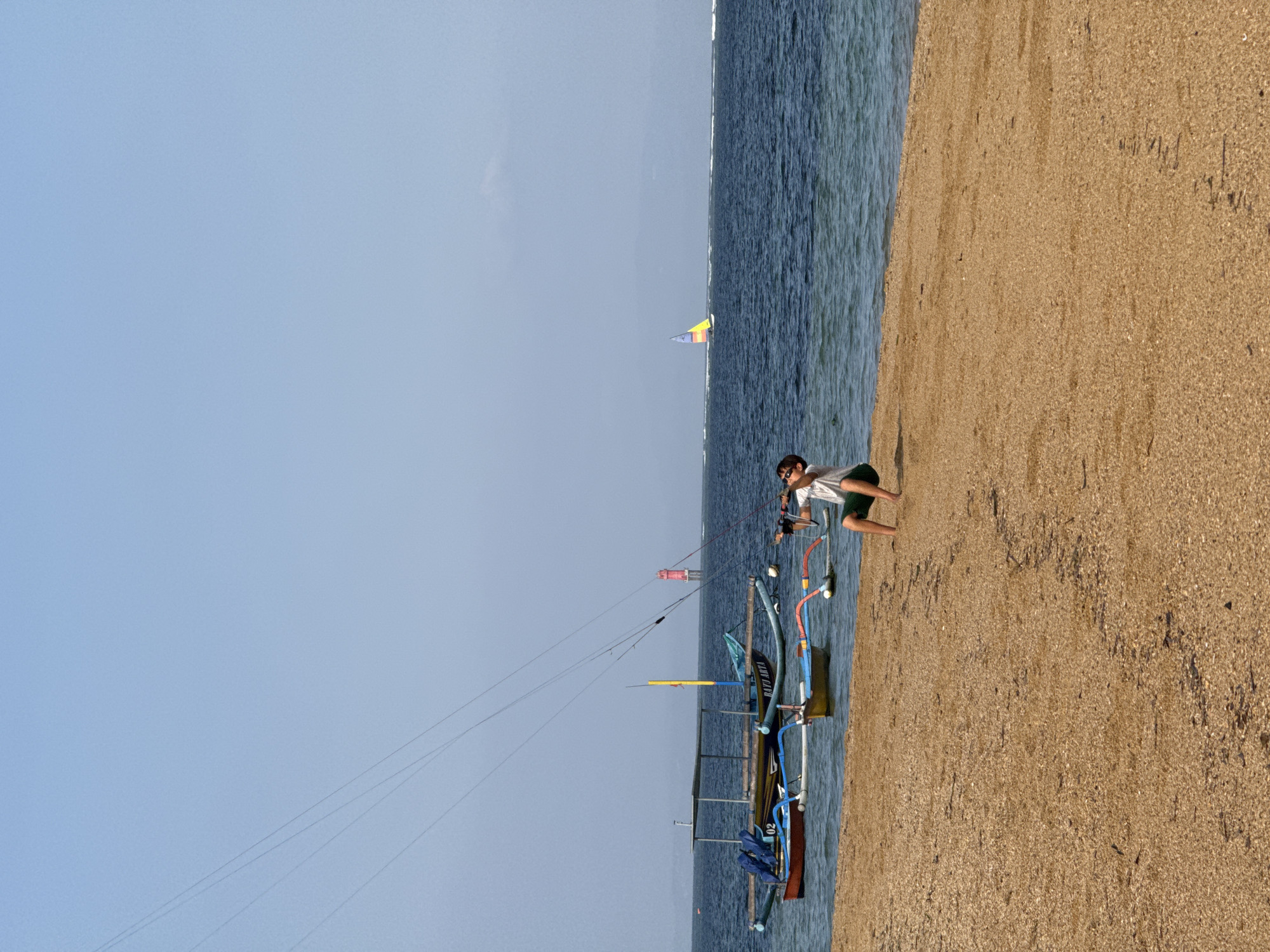 The image depicts a beachfront scene at Sanur Beach, Denpasar, Indonesia. A person is shown on the sand, holding a kiteboarding control bar, appearing to be in the process of launching or controlling a kite, as indicated by the lines extending upwards and out of frame. The person is wearing a white t-shirt and green shorts, with sunglasses on. In the water, several traditional Balinese jukung boats are anchored near the shore. Further out in the ocean, a red lighthouse is visible, and a small sailboat with colorful sails is sailing in the distance. The sky is clear and blue, and the water is a deep blue color. The beach itself is sandy, with some small debris visible.
