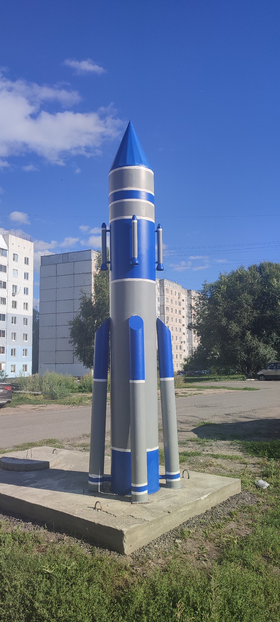 A large, gray and blue, cylindrical monument resembling a rocket stands on a concrete foundation in a public space. The monument has a pointed blue top with white trim, and several cylindrical elements protrude from its sides. The rocket is surrounded by grass and sits in front of apartment buildings. In the background, trees and parked cars are visible. The sky is blue with some white clouds. The monument is located in the city of Novoaltaysk, Russia, likely in a residential area given the presence of apartment buildings and a paved road. No people are visible in the image.