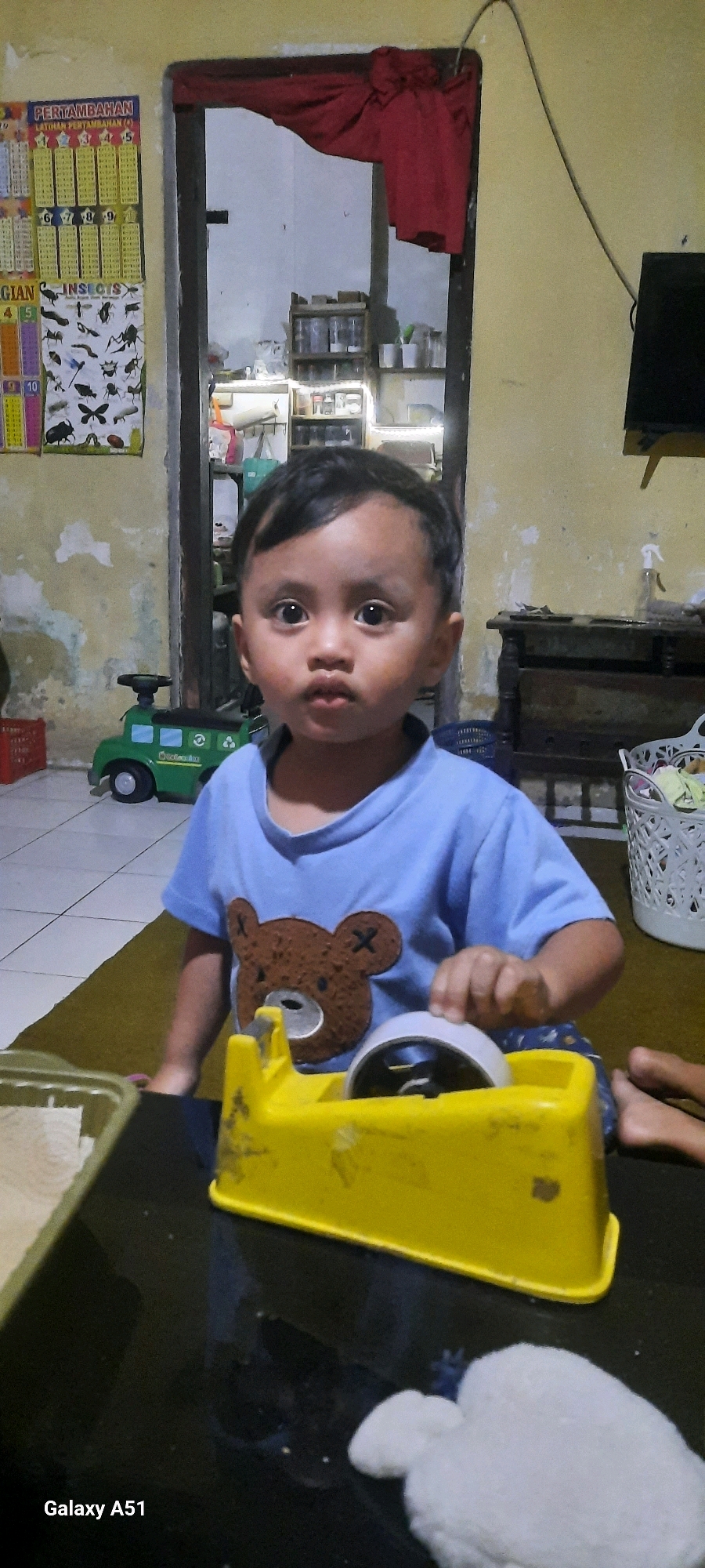 A young child, appearing to be of Southeast Asian descent, is the primary subject of this indoor photograph, likely taken in Jatitengah, Indonesia. The child is looking directly at the camera with wide eyes, exhibiting a neutral expression. They are wearing a light blue t-shirt with a brown bear graphic on the front. The child is seated at a dark-colored table, actively engaging with a bright yellow tape dispenser.
The background reveals a home environment. To the left of the child, a green toy truck is visible on the floor, and further left, a poster with Indonesian text and images of insects is affixed to a wall. Behind the child, a doorway leads into another room where shelves stocked with various items, possibly jars and bottles, are illuminated. A red cloth hangs over the top of the doorway. To the right of the child, part of a black television screen is visible, mounted on a stand. Another white basket is also present to the right, containing some fabric. The walls appear to be painted a light yellow and show signs of wear. The lighting in the room is subdued, suggesting it may be evening or a dimly lit interior. The "Galaxy A51" watermark in the lower left corner indicates the photograph was taken with a Samsung Galaxy A51 smartphone.