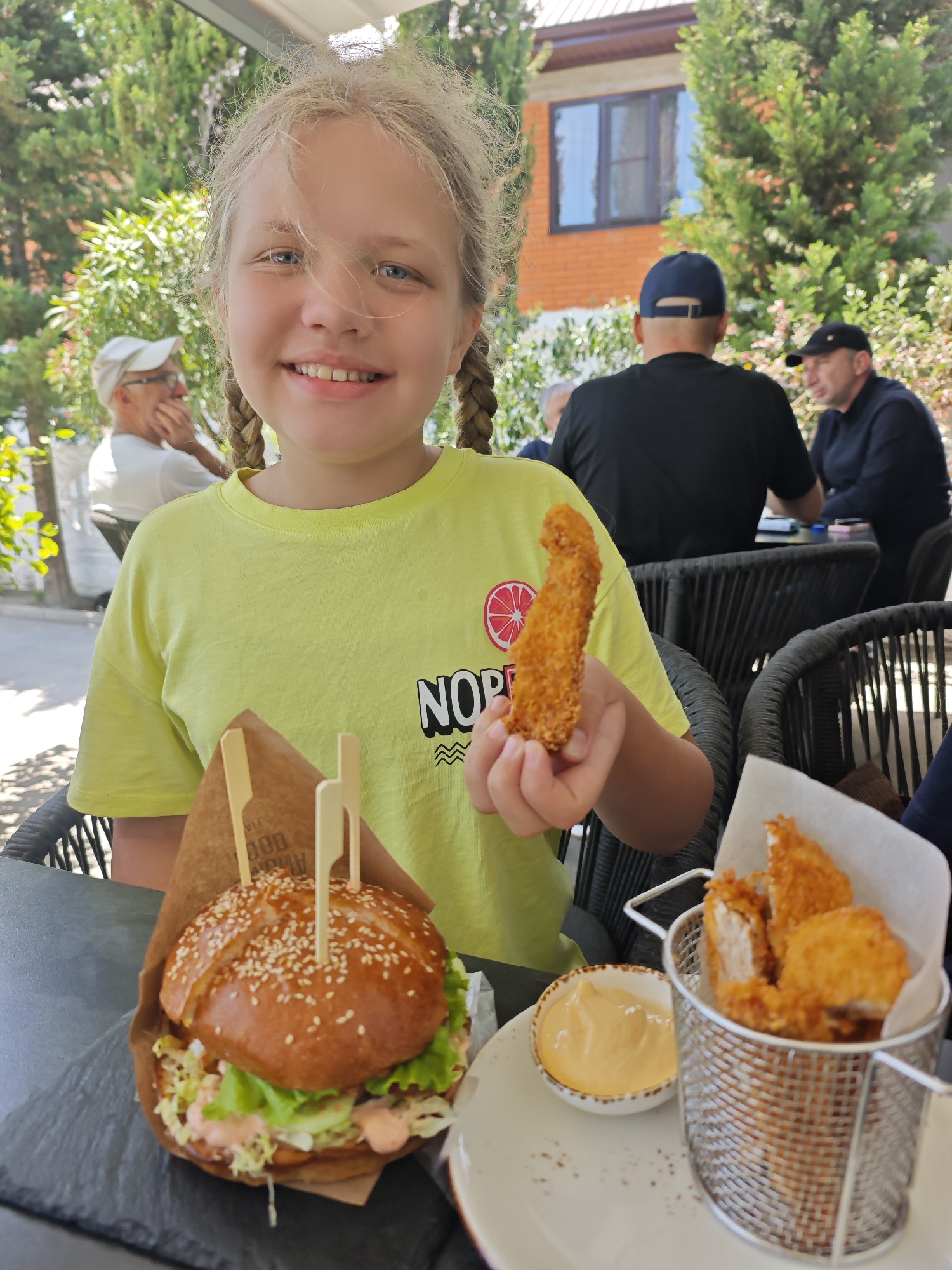 A young girl with blonde hair, styled in braids and partially obscuring her face, smiles directly at the camera while seated at an outdoor cafe table in Gudauta, Abkhazia. She wears a neon green t-shirt with a grapefruit graphic and partial text "NOR." In her right hand, she holds a golden-brown, breaded chicken tender. On the dark table in front of her, a large burger with a sesame seed bun, lettuce, and sauce, secured by two wooden picks, sits within a brown paper wrapper. Adjacent to the burger is a small metal basket containing several more fried chicken pieces, accompanied by a small bowl of light orange dipping sauce. In the background, several male patrons are seated at other tables; an older man in a white cap is visible to the left, and two men in dark caps are visible to the right, one with his back to the camera. Lush green trees and bushes surround the outdoor seating area, with a brick building featuring a dark-framed window visible further back. The scene is brightly lit by natural daylight.