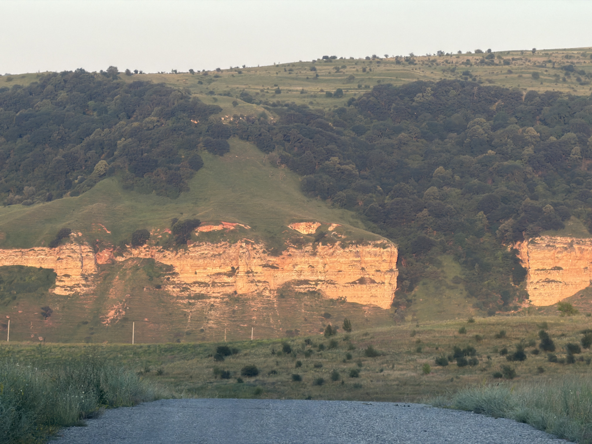 A long shot captures a gravel road leading towards a vast, treeless plain and a towering cliff face illuminated by the warm hues of sunset. The cliff, with its distinct layers of orange and brown rock, is the focal point of the midground, casting long shadows across the grassy slopes that ascend behind it. Dense forest blankets the upper reaches of the hills, creating a stark contrast with the barrenness of the cliff and the open plain below. The sky above is a pale, washed-out blue, typical of early evening. The foreground is dominated by the gravel road and the tall, dry grass that lines its edge. There are no people or animals visible in the scene.