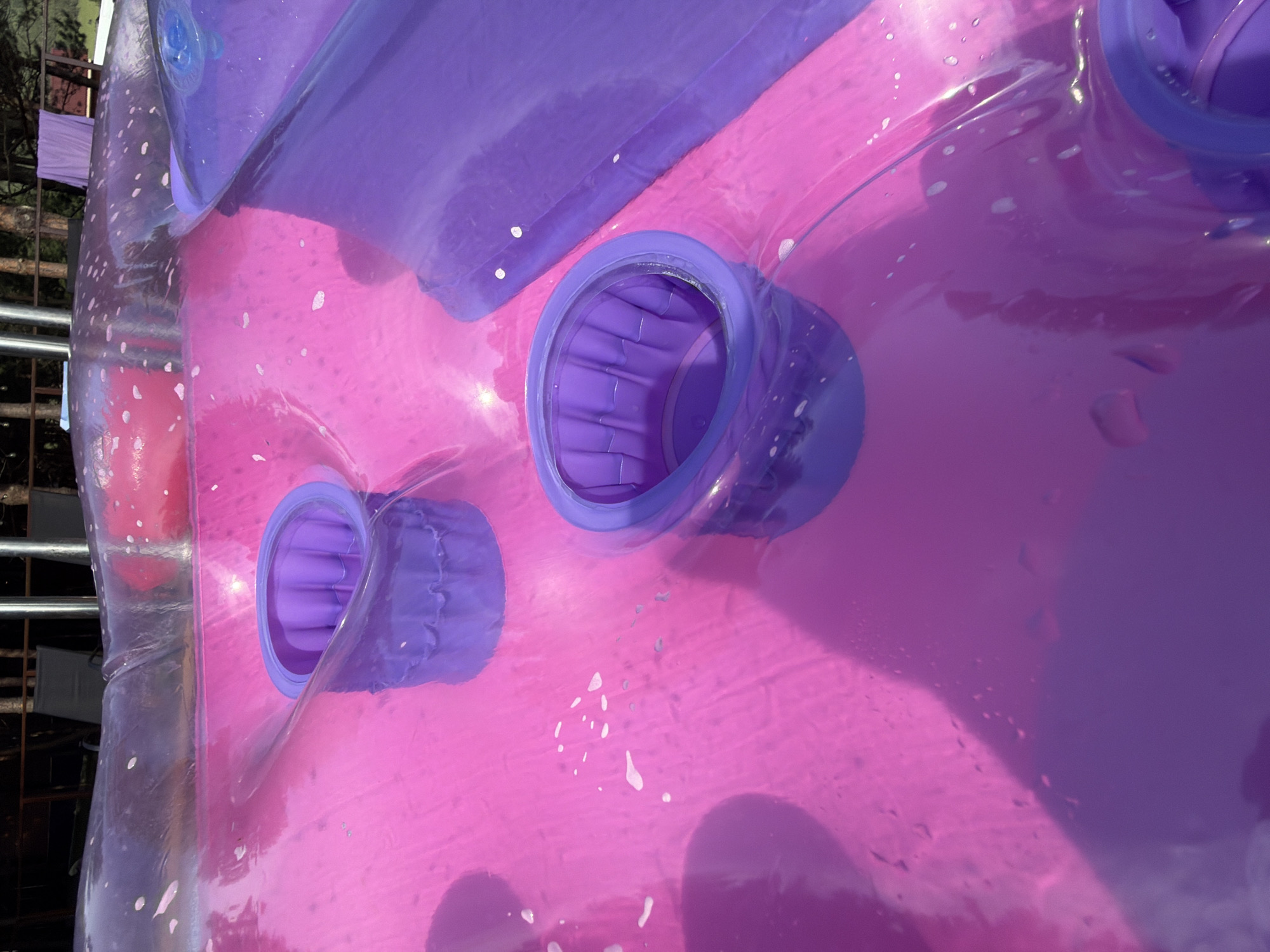 The media file displays a close-up, angled shot of a pink and purple inflatable structure. The surface appears to be made of a translucent, glossy plastic, possibly PVC, and is speckled with small, white water droplets or air bubbles. Two prominent purple, cylindrical openings are visible on the surface, each with a ridged interior. These openings are set into the pink material. In the background, a metal railing and some foliage are slightly visible, suggesting an outdoor setting. The lighting suggests it is daytime, with some shadows and reflections indicating bright conditions. The overall impression is of a playful, possibly recreational item, given its inflatable nature and vibrant colors. The image is abstract, focusing on the texture and form of the inflatable. There is no visible text within the frame. The location is identified as Elekmonar, Russia.