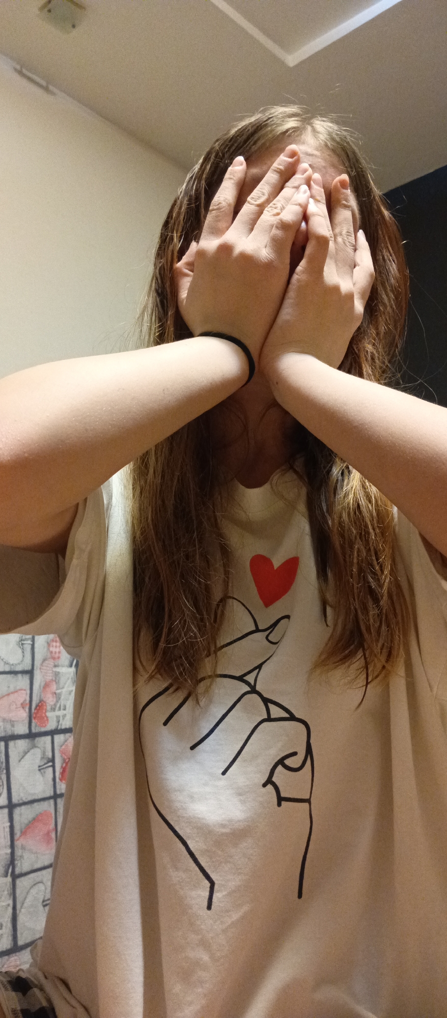 A person stands indoors, concealing their face with their hands. They are wearing a light-colored t-shirt adorned with a graphic of a hand making a heart gesture, accompanied by a red heart above the hand. The person's arms are visible, with a black elastic band around their left wrist. Their long, brown hair partially obscures the t-shirt design. The background consists of a light-colored wall, a darker wall to the right, and a glimpse of patterned material to the left. The location is described as being in the city of Unknown, Unknown.
