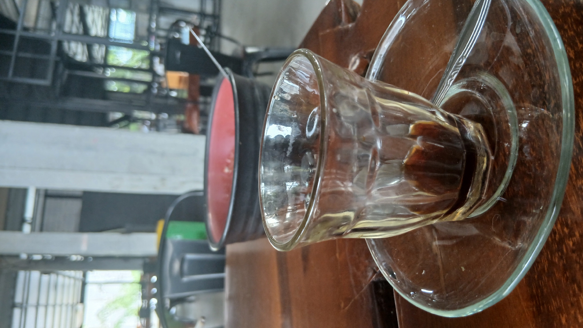 A close-up, eye-level shot focuses on a small, clear glass tumbler containing a dark liquid, likely coffee, with a minimal amount remaining at the bottom. The glass sits on a clear glass saucer, which rests on a dark, polished wooden table. To the left of the glass, the edge of a spoon is visible on the saucer. In the background, slightly out of focus, a large black bowl with a red interior is visible, with a metal spoon protruding from it. Further back, the interior of an establishment is apparent, with concrete pillars, metal framing, and seating areas suggesting a cafe or restaurant setting. Natural light filters in from windows or openings in the background. The overall impression is a still life of a drink on a table within a public space.