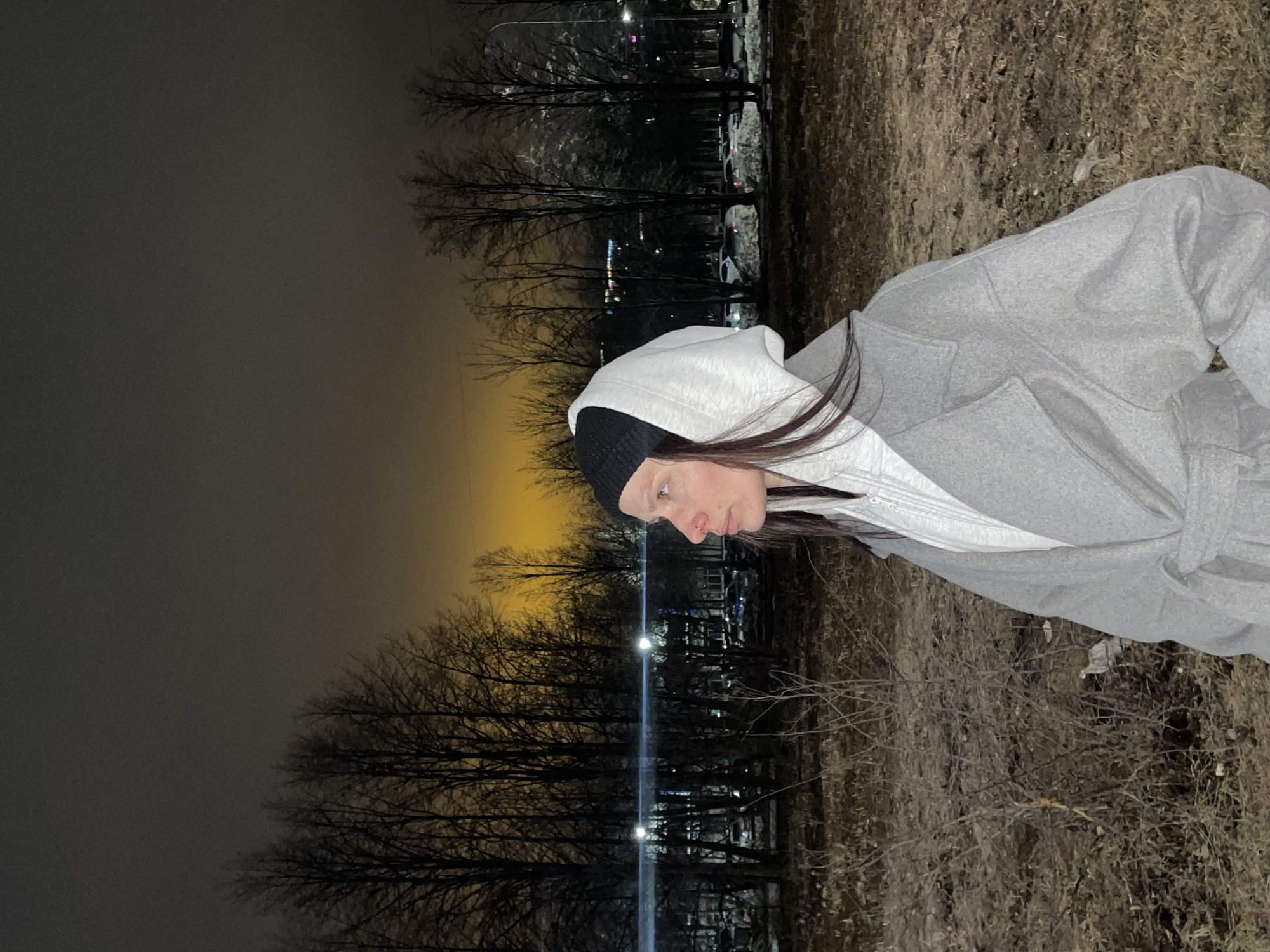 A woman wearing a gray coat with a hood and a black beanie stands outside at night. The location appears to be a park or wooded area in Izhevsk, Russia, with bare trees and sparse vegetation. In the background, city lights and parked cars are visible. The sky is dark and cloudy.