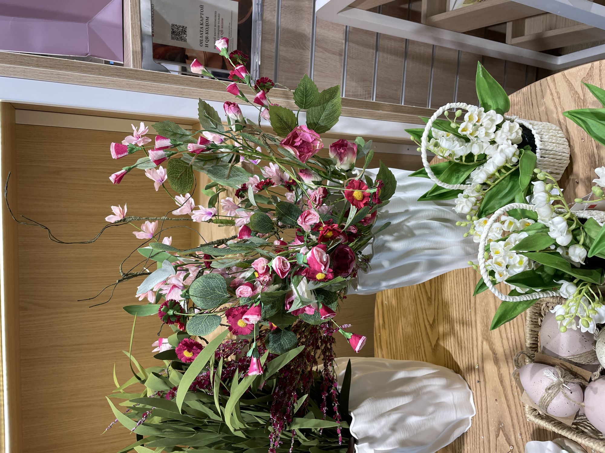 Here is a description of the image:
The image shows a collection of floral arrangements indoors. There are two vases filled with flowers, one with pink and burgundy blooms and another with greenery and purple accents, draped with white fabric. Next to these, there are two baskets containing white lily of the valley arrangements. Easter-themed decorated eggs are also visible in one of the baskets. The setting appears to be a store or home interior, with wooden shelves and surfaces visible in the background. A price tag and a sign that says "Payment by card and QR code possible only for some items via QR code" in Russian are partially visible. The image seems to be taken at an angle, looking down at the floral displays.