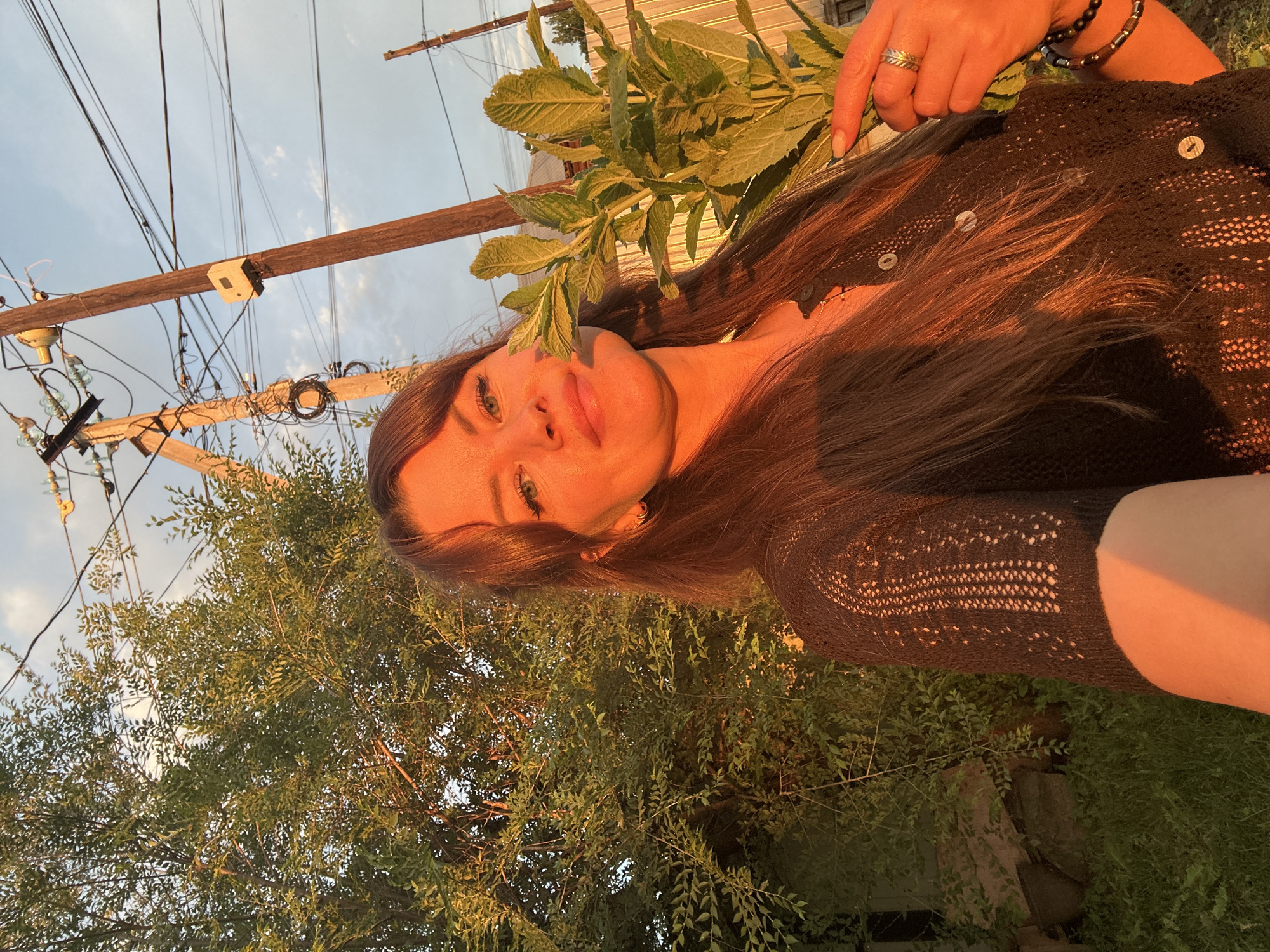 A woman with long, dark brown hair and green eyes is captured in a selfie outdoors, bathed in the warm, golden light of what appears to be sunset. She is smiling slightly and holding a bunch of fresh green mint in her right hand. The woman is wearing a black, loosely knitted cardigan with buttons. Her left hand is visible, adorned with rings and bracelets.
The background features lush green foliage from a tree and shrubbery to her left, and a light-colored wooden fence to her right. In the distance, a utility pole with numerous electrical wires and insulators is prominent against a blue sky with scattered clouds. The overall impression is one of a serene moment captured during the golden hour, possibly in a residential area of Chaikovsky, Russia, given the presence of the utility infrastructure and the style of fencing.