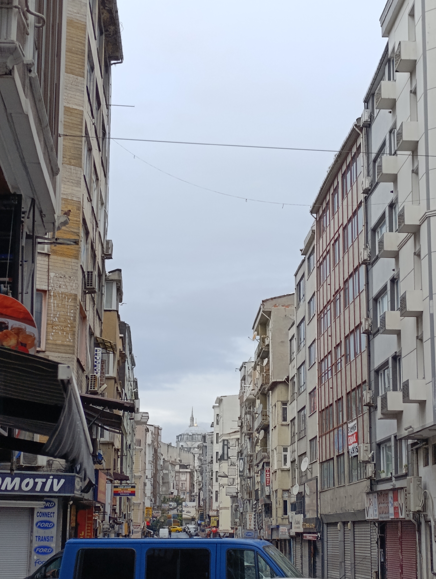 The image captures a narrow street scene in Istanbul, Turkey. Tall buildings line both sides of the street, creating a deep perspective towards the city center where a mosque dome is visible in the distance. Utility cables crisscross overhead. Storefronts and awnings are visible on the lower levels of the buildings. A blue van occupies the foreground, obscuring part of the street and storefronts. There are indications of pedestrian and vehicular traffic in the middle distance. The sky is overcast.