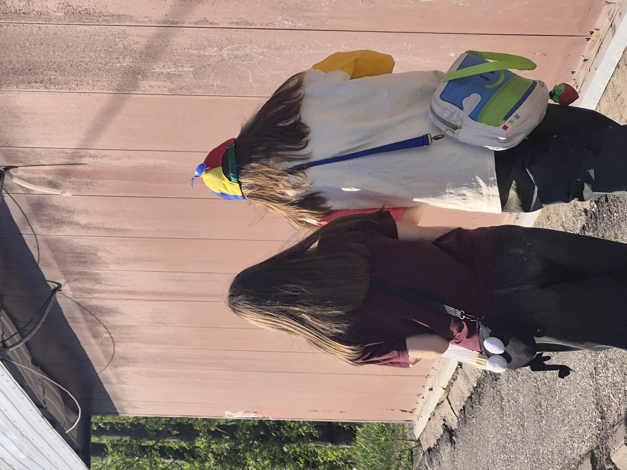 Two individuals are standing on a paved surface, likely a sidewalk, adjacent to a light-colored, horizontal plank wall. The lower portion of the scene includes green foliage, possibly trees or shrubs.
The person in the foreground is wearing a maroon shirt and black pants. They have long, dark hair that extends down their back. They are carrying a black bag or purse with white embellishments and a dark strap over their shoulder.
The individual in the background is wearing a white shirt and black pants. They have long brown hair and are wearing a colorful, striped hat. They are also carrying a white bag with blue and green accents, suspended from a blue strap. A small red strawberry toy is attached to the bag. A yellow fabric item is visible protruding from beneath the white shirt.
The location is indeterminate.