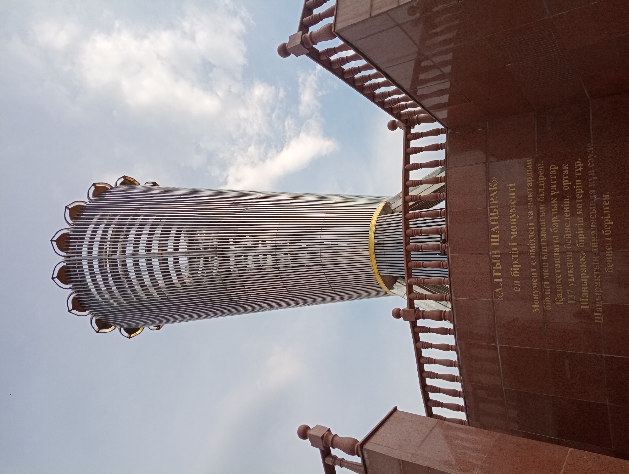 This low-angle, upward-looking shot captures the "Golden Shanyrak" (Golden Skylight) monument, a significant landmark in Shymkent, Kazakhstan. The monument, a tall, cylindrical structure with a ribbed, metallic exterior, is prominently featured against a backdrop of a partly cloudy sky, suggesting it is daytime. The top of the structure is adorned with petal-like elements, hinting at a stylized representation. A golden band encircles the monument partway up.
To the right, a reddish-brown marble wall serves as a base or part of the monument's surroundings. This wall features ornate wooden railings with turned balusters and finials, adding a traditional architectural element. Engraved in gold lettering on the marble wall is text in Kazakh. The text, transliterated and translated, reads: "«ALTYN SHANYRAQ» monument of national unity. The monument embodies the unity and solidarity of the peoples of our country. All the nations in Kazakhstan are depicted as 137 arrows, united and rising to the common shanyrak. Around the shanyrak, the sun's rays are depicted." This inscription provides crucial context about the monument's symbolic meaning, representing the unity of Kazakhstan's diverse ethnic groups. The overall impression is one of grandeur and national pride, conveyed through the monument's imposing design and the inscription's message of unity.