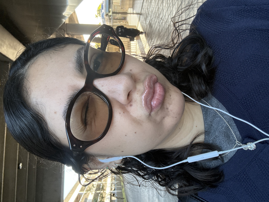 A front-facing, waist-up image captures an adult female with dark, wavy hair, illuminated by natural light in an outdoor urban setting. She wears tortoise-shell oval sunglasses with light amber lenses; her right eye is visibly closed in a wink, creating wrinkles at the outer corner. Her lips are pursed, appearing glossy with a light pink or nude product. White wired earphones are in both ears, with an inline control visible on the wire resting on her chest. Her right ear features multiple piercings, including a stud with a green stone and several silver rings. She is dressed in a dark blue v-neck sweater over a grey shirt. A silver ball-chain necklace with a circular, clear faceted pendant hangs around her neck. In the background, a large, grey concrete elevated structure dominates the upper frame. To the left, a multi-story building is partially visible. To the right, a blurry, dark silhouette of another individual walks away behind a vertical-barred metal fence. The environment is consistent with urban infrastructure, such as elevated transit lines or roadways, within the city of Hialeah, Florida, United States of America.
