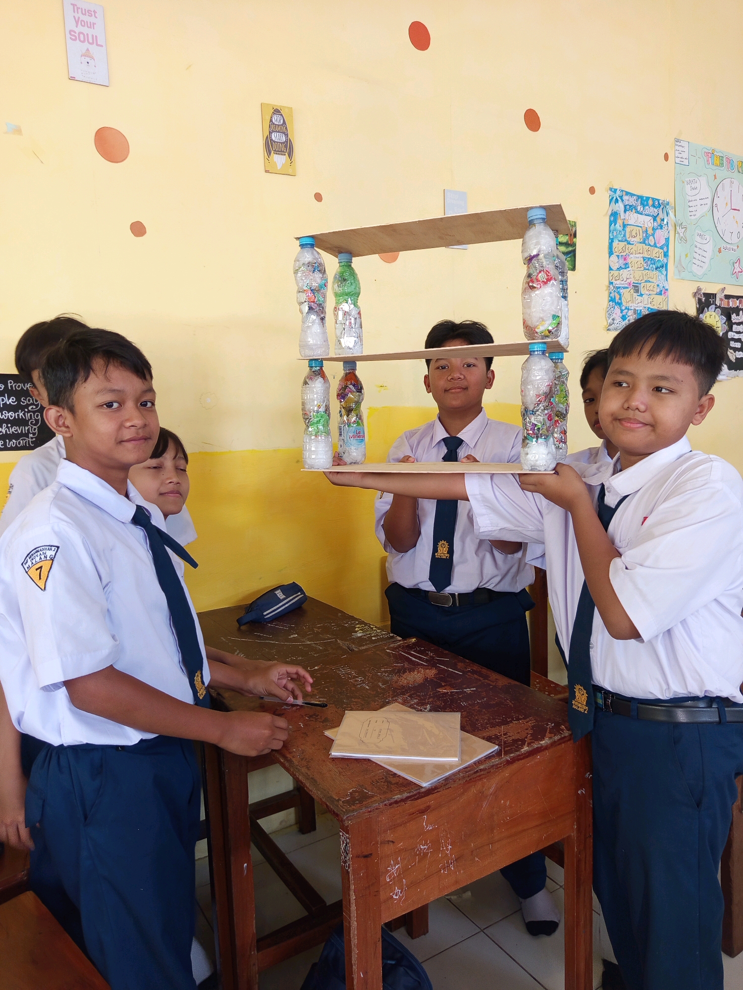 The image shows five male students and one female student, all appearing to be of elementary or middle school age, in a classroom. The students are wearing school uniforms consisting of white shirts and dark blue pants or skirts, with ties. The students are gathered around a wooden desk, some holding a makeshift shelf constructed from wood and plastic bottles. The shelf is filled with plastic bottles. The classroom walls are painted yellow and decorated with various posters and drawings. One poster, seen in the upper left, reads "Trust Your SOUL". The room appears to be illuminated by natural light. The students' uniforms suggest they are students at a school in Malang, Indonesia, as indicated by the emblem on the shirt of the student in the foreground, which displays the text "YAYASAN MADRASAH ALIYAH MALANG 7". This emblem identifies the school as being part of an educational foundation in Malang.