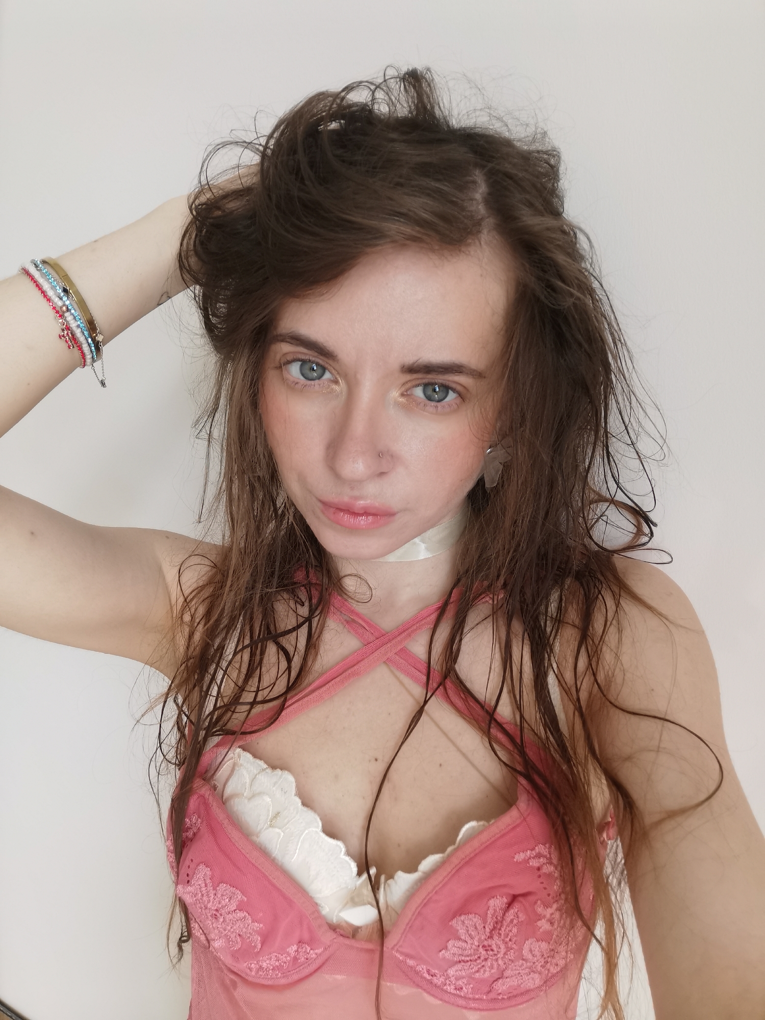A woman poses for a selfie against a white wall. She is wearing a pink and white lingerie top and a white ribbon around her neck. Her brown hair is styled messily, with some strands appearing wet. She has blue eyes and a nose piercing. Her left arm is raised, with bracelets visible on her wrist. The background is plain and white. The location is not discernible in the image.