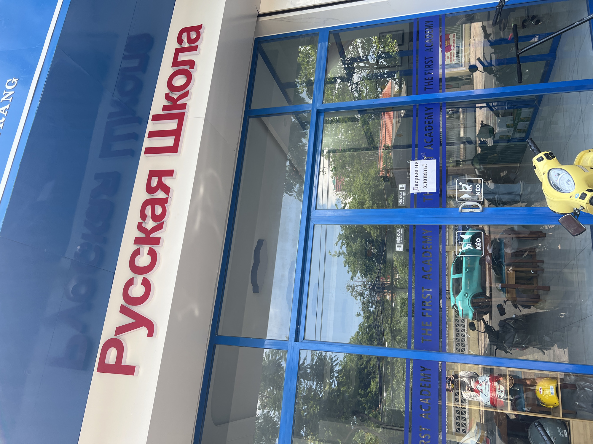 The image shows the storefront of "The First Academy" in Bac Nha Trang Ward, Vietnam. The signage includes the words "Русская Школа" (Russian School) in red lettering. A yellow scooter is parked in front of the glass doors, reflecting the surrounding environment. Stickers with Vietnamese and Russian instructions about the door (possibly "Slide to Open" and "Do not slam the door!") are visible on the glass. Inside, a light blue car and other objects are reflected in the glass.