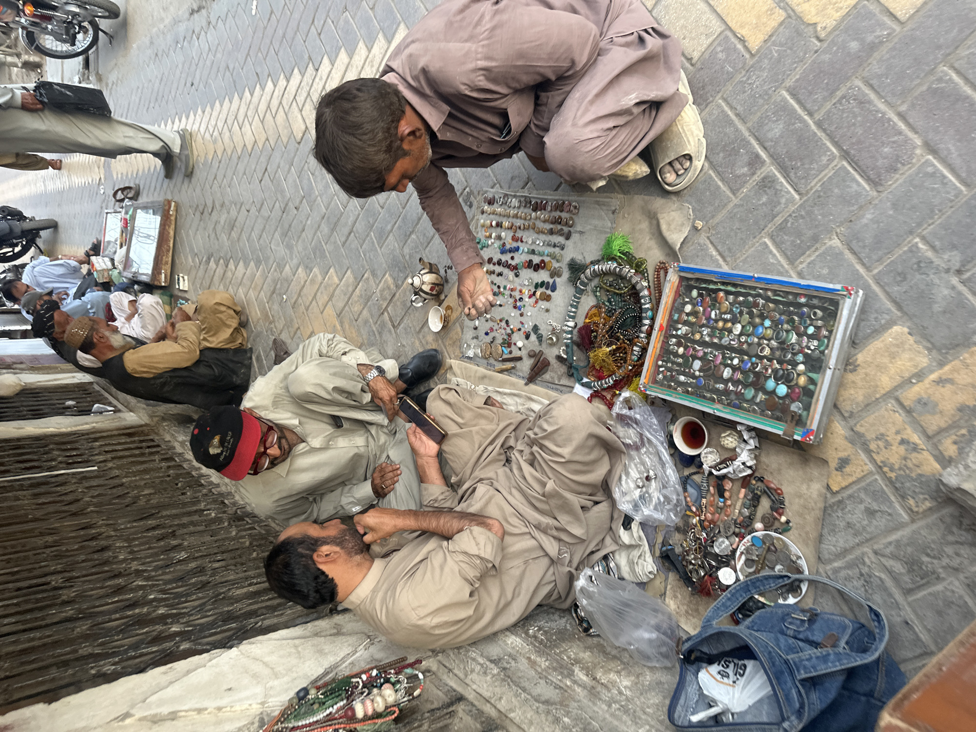 This is a street scene in Quetta City Tehsil, Pakistan, showing several men engaged in commerce. In the foreground, two men are seated on the ground, surrounded by various items for sale. One man, wearing a light-colored tunic and pants, is holding a smartphone and appears to be showing it to the other man, who is wearing a darker tunic and pants. The second man is crouched and examining a collection of colorful gemstones laid out on a cloth. Several strings of beads and jewelry are also visible around them.
Further back, other men are seated or standing, with some appearing to be customers or vendors. One man in a light-colored outfit is sitting near a metal grate, and another is seen from behind wearing a light blue outfit. Items on display include jewelry, gemstones, and possibly other small goods. Motorcycles are parked on the street in the background, indicating a busy urban environment. The ground is paved with brick-like tiles. The overall impression is of a bustling marketplace.