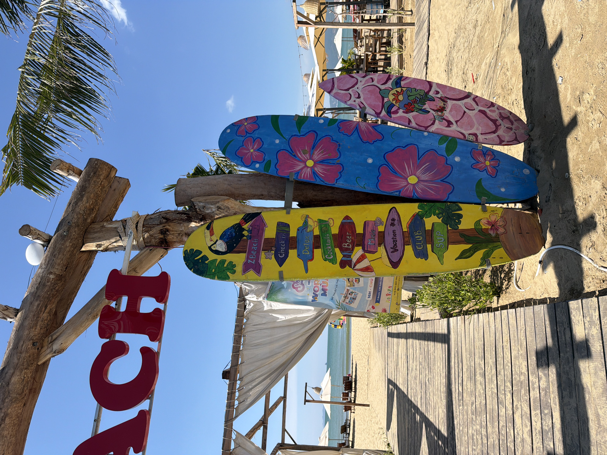 This is a medium shot photograph of a beach entrance in Quy Nhon Ward, Vietnam, on a sunny day. The foreground features a wooden boardwalk leading towards the beach. To the right of the boardwalk, leaning against a rustic wooden structure, are three colorful surfboards. The left-most surfboard is yellow and features various directional arrows pointing to "Zbeach," "Check In," "Detox," "Cocktails," "Beer," "Music," "Vacation," "Club," and "Sup." It also has a painted toucan and tropical leaves. The middle surfboard is blue with large pink hibiscus flowers and a smaller pink flower. The right-most surfboard is light pink with a white honeycomb-like pattern and a drawing of a sea turtle wearing sunglasses.
Behind the surfboards, a white canopy with draped curtains offers shade over a beach seating area. Further back, beach chairs and umbrellas are visible on the sandy beach, with the calm ocean stretching to the horizon under a clear blue sky. A palm frond hangs from the top right of the frame. The sign above the surfboards reads "BEACH" in large red letters. The overall scene depicts a relaxed, tropical beach environment.