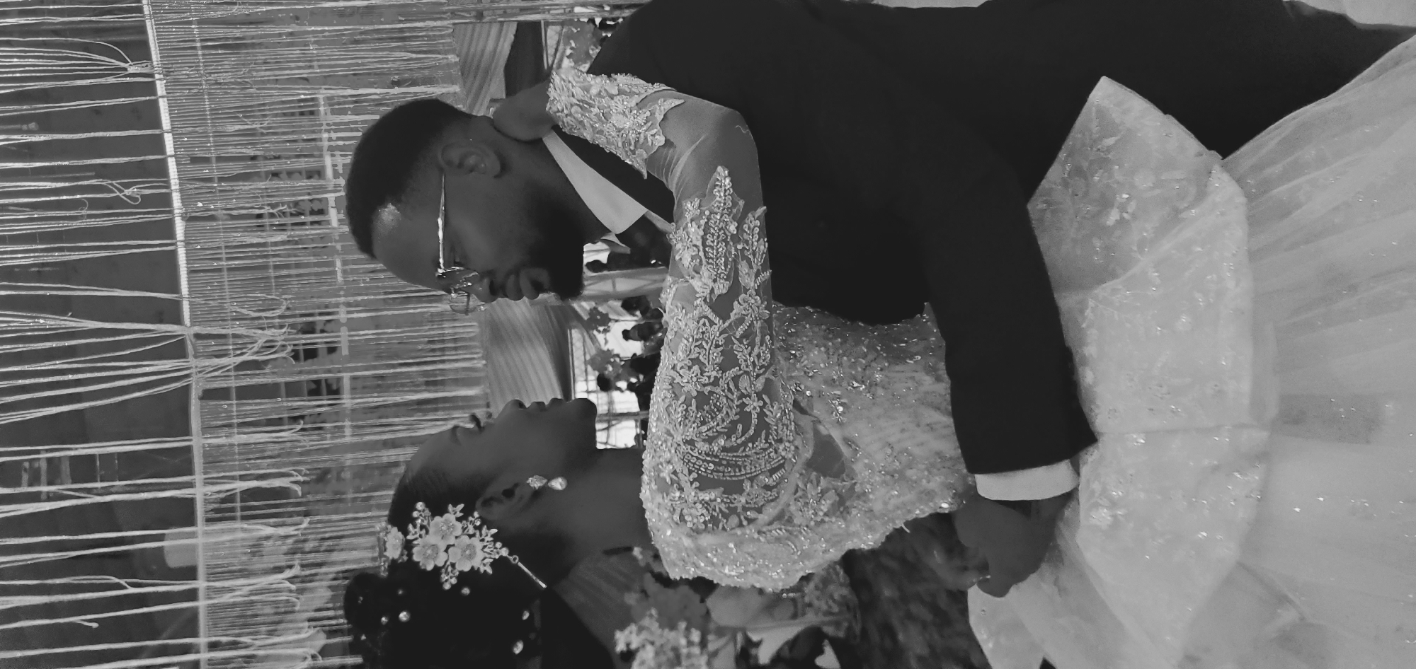 A black and white photograph depicts two individuals, a female and a male, in a close embrace within a decorated indoor venue.
The female, positioned on the left, has dark hair styled upwards, adorned with a floral and beaded hairpiece. She wears drop earrings and a light-colored, long-sleeved gown featuring intricate lace patterns and beadwork on the bodice and sleeves. Her left hand is visible near the male's lower torso, and her right arm is around his neck/shoulder. Her profile is visible, with her gaze directed towards the male.
The male, positioned on the right, has dark, neatly trimmed hair and a beard. He wears clear-framed glasses, a dark suit, a white shirt, and a dark tie. His arms are around the female's waist/lower back. His profile is visible, with his gaze directed towards the female.
The background features numerous thin, light-colored vertical strands hanging from above, suggesting decorative elements in an event space. Other indistinct shapes and ambient lighting are present in the distant background. The specific location in "Lankan, Nigeria" is not discernible from the visual information presented in the image.